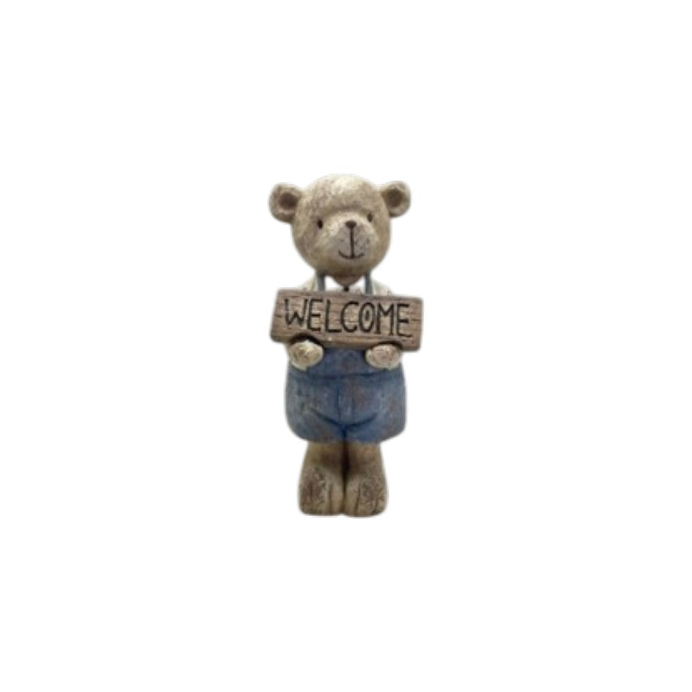 A small decorative teddy bear figurine holding a rectangular sign that reads “WELCOME.” The bear has a weathered beige/tan face and body with blue pants, shown against a plain white background.