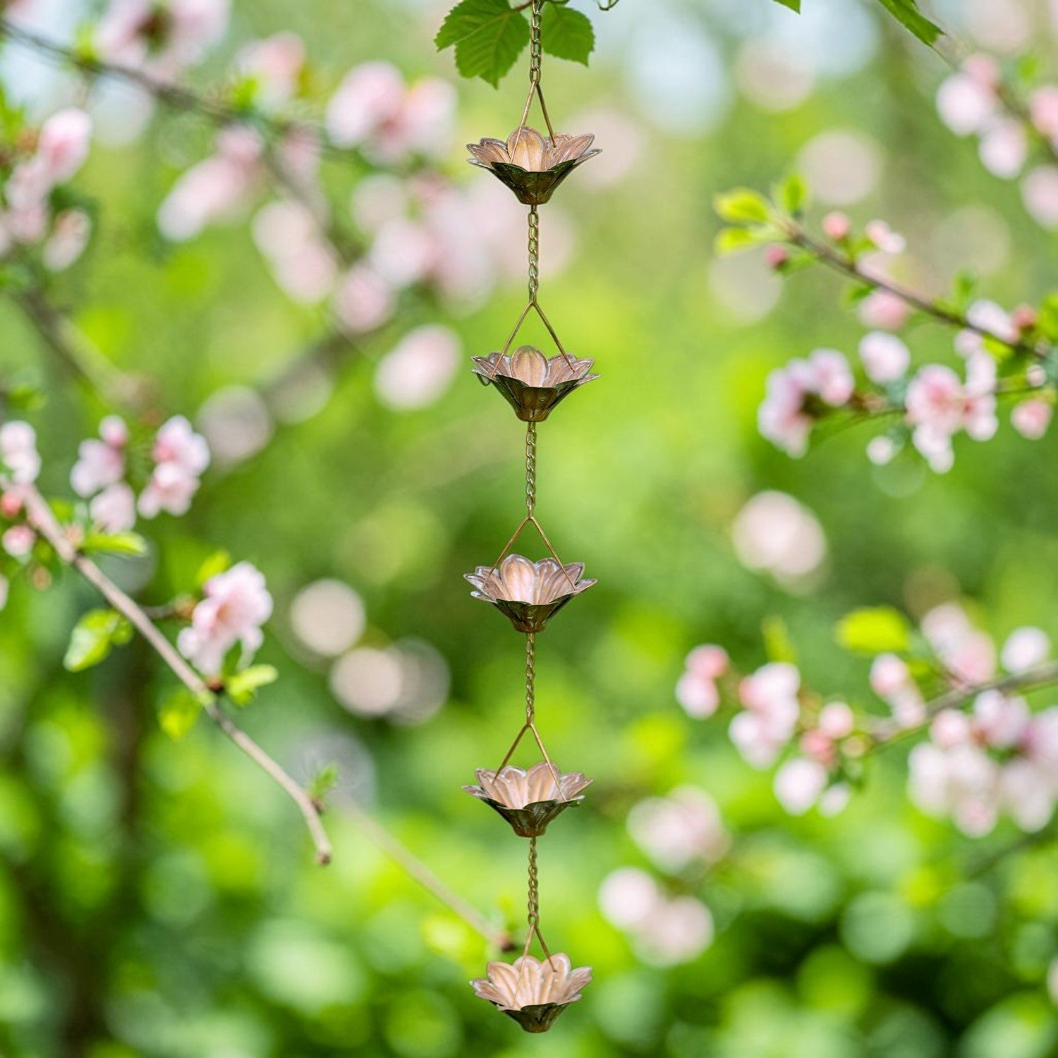 A hanging flower wind chime with five metallic lotus-shaped bells in soft pink and bronze tones, suspended by a chain in a garden with green leaves and pink blossoms in the background.