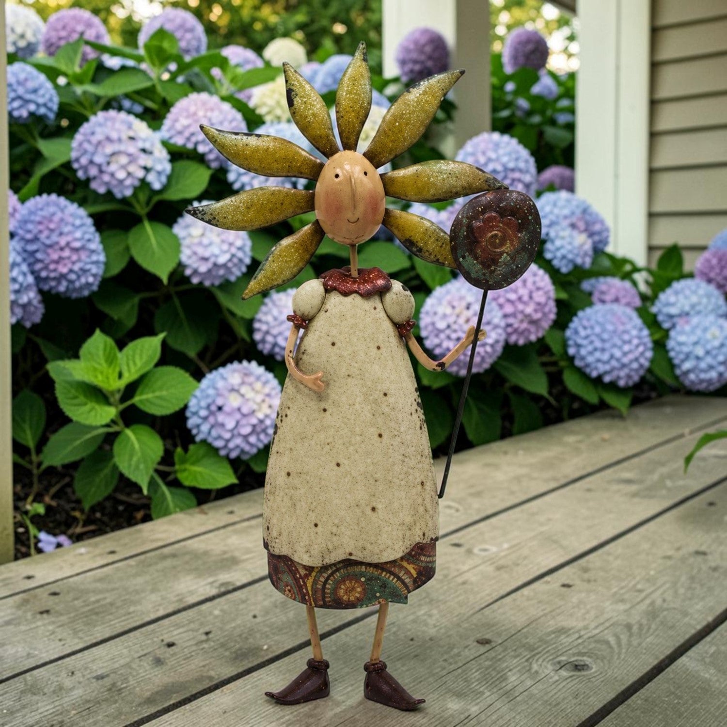 A whimsical metal garden figurine shaped like a sunflower-headed woman in a dress, standing on a wooden deck with purple hydrangeas in the background.