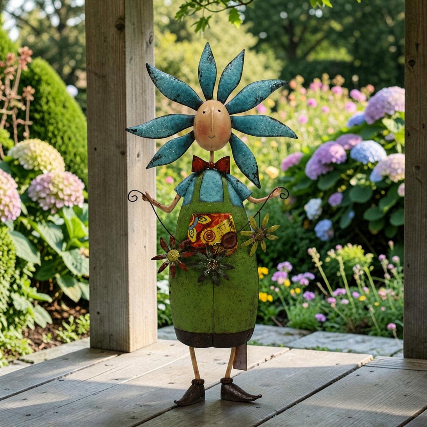A whimsical metal garden figurine shaped like a flower-headed character standing on a wooden deck in a lush garden. The figure has a blue flower head, a green body, a red bow tie, and floral details on its torso.