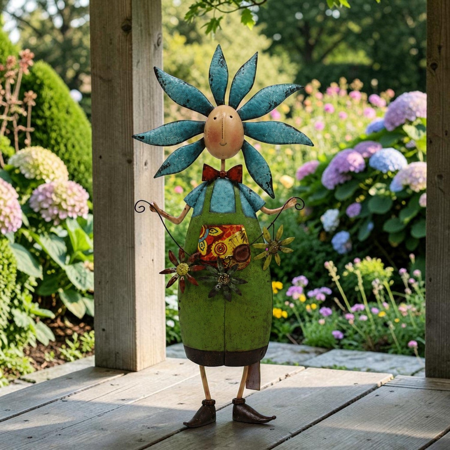 A whimsical metal garden figurine shaped like a flower-headed character standing on a wooden deck in a lush garden. The figure has a blue flower head, a green body, a red bow tie, and floral details on its torso.