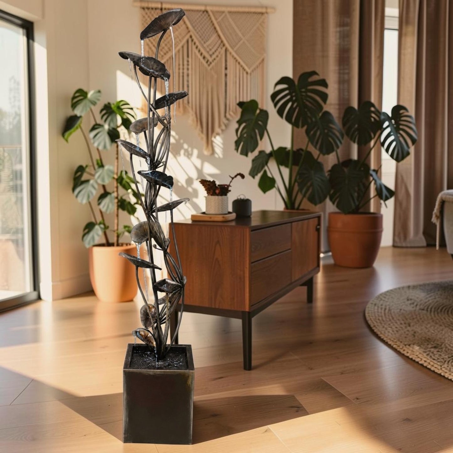 A tall decorative metal plant sculpture with dark leaf-like branches in a square dark planter, placed in a sunlit modern living room with wood floors, potted plants, and a wooden sideboard.