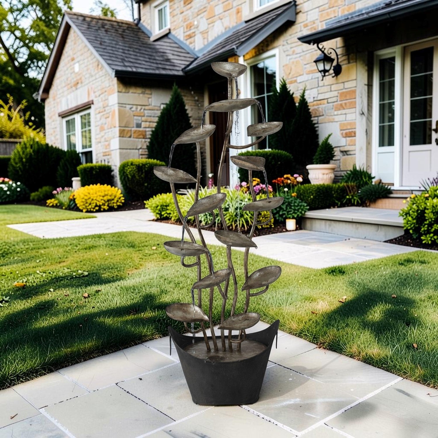 A modern abstract metal garden sculpture with layered leaf-like discs on a dark base, displayed on a stone patio in a landscaped front yard in front of a stone house.