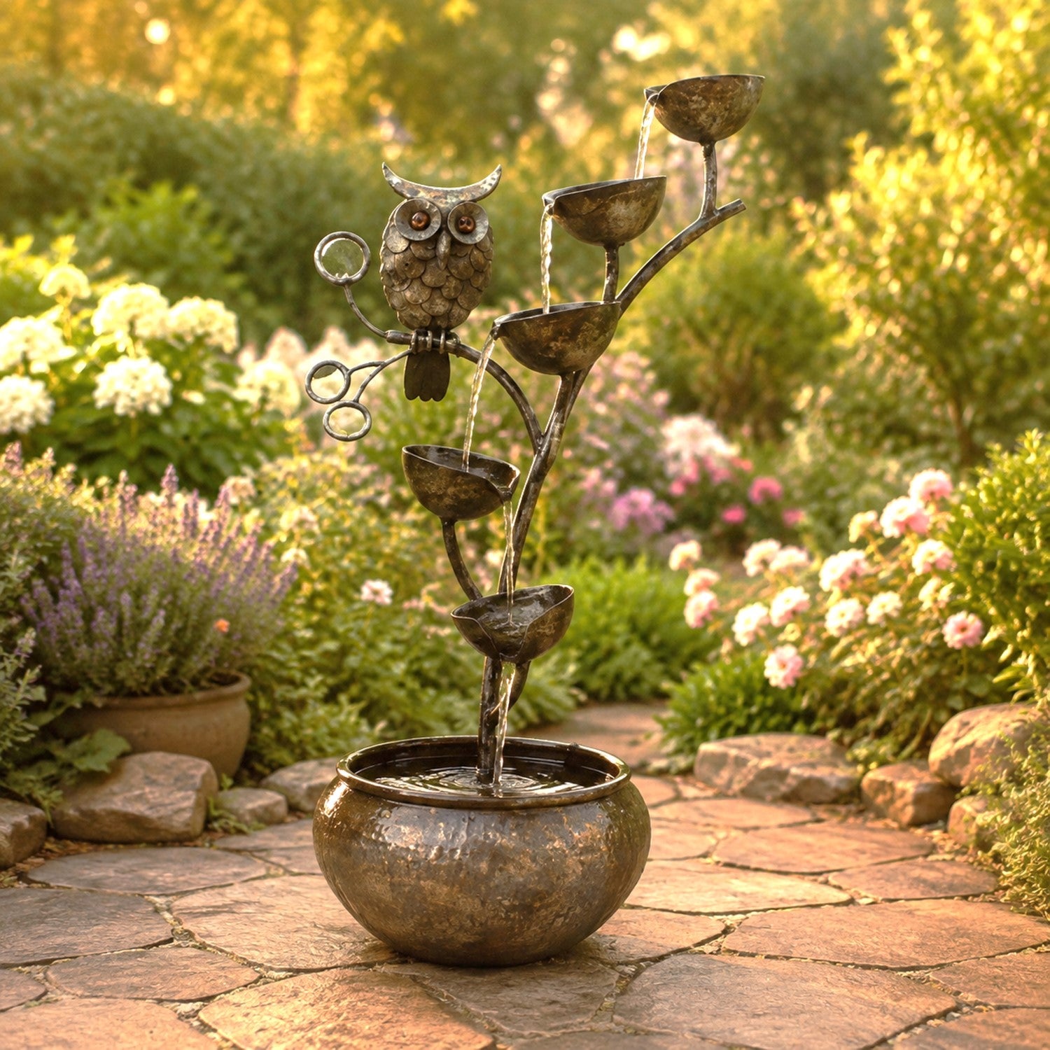 Metal garden fountain sculpture featuring an owl perched on a branch-like structure with multiple tiers of bowls from which water flows into a round basin, set on a stone path in a landscaped garden.