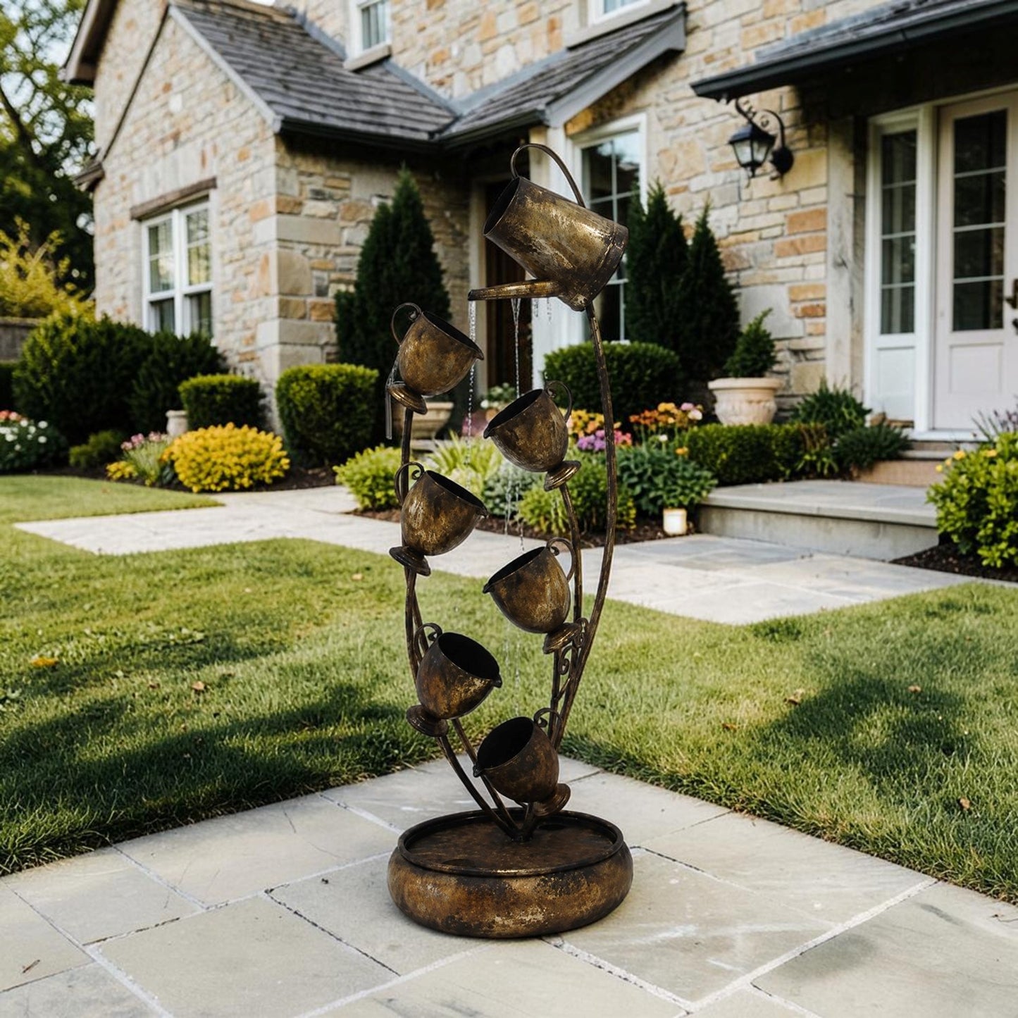A rustic metal garden fountain sculpture with multiple hanging cups and a circular base, displayed outdoors on a stone patio in front of a stone house with a green lawn.