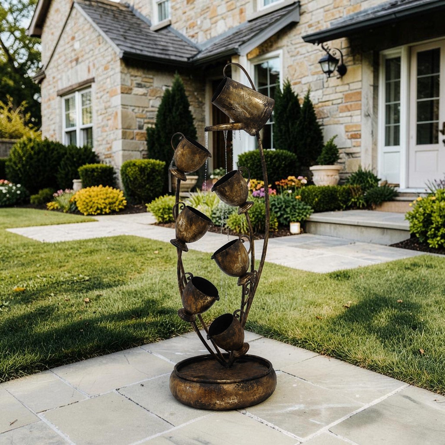 A rustic metal garden fountain sculpture with multiple hanging cups and a circular base, displayed outdoors on a stone patio in front of a stone house with a green lawn.