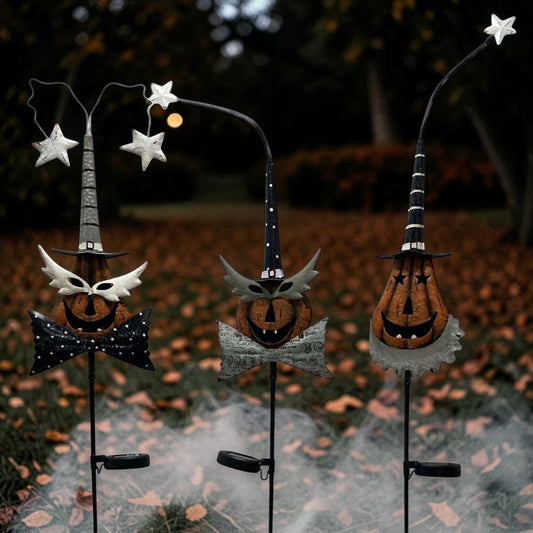 Three Halloween yard stake decorations displayed outdoors on a leaf-covered ground with mist. The decorations include witch hats, jack-o'-lantern faces, bows, and silver stars on curved black metal stakes.