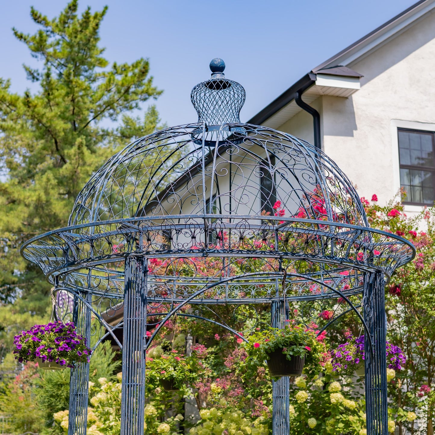 A large metal garden gazebo with a blue-gray painted frame and intricate scrollwork, decorated with hanging flower baskets and surrounded by blooming pink and purple flowers in a garden setting.