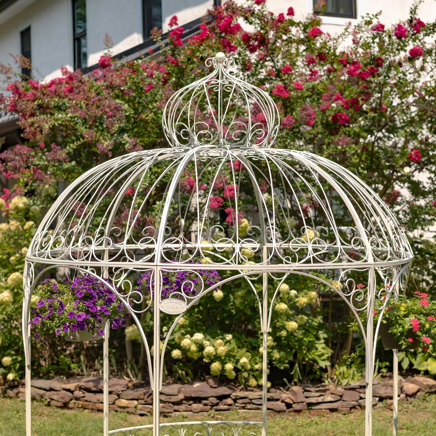 Decorative white metal gazebo in a garden with flowers and a house in the background