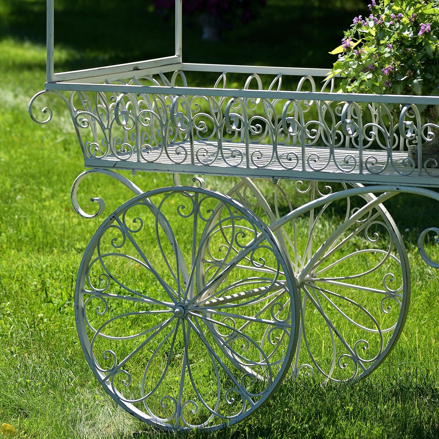 A decorative white wrought-iron garden cart with ornate scrollwork and large spoked wheels, photographed outdoors on a green lawn with flowers in the upper right corner.
