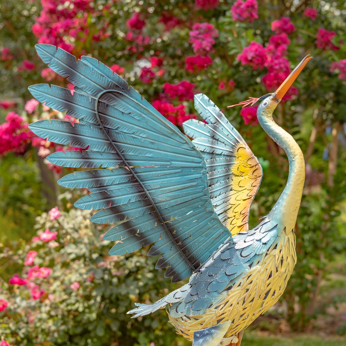 A decorative metal garden statue of a heron with wings spread, featuring a blue and yellow patterned body, set against a blurred background of green foliage and pink flowers.