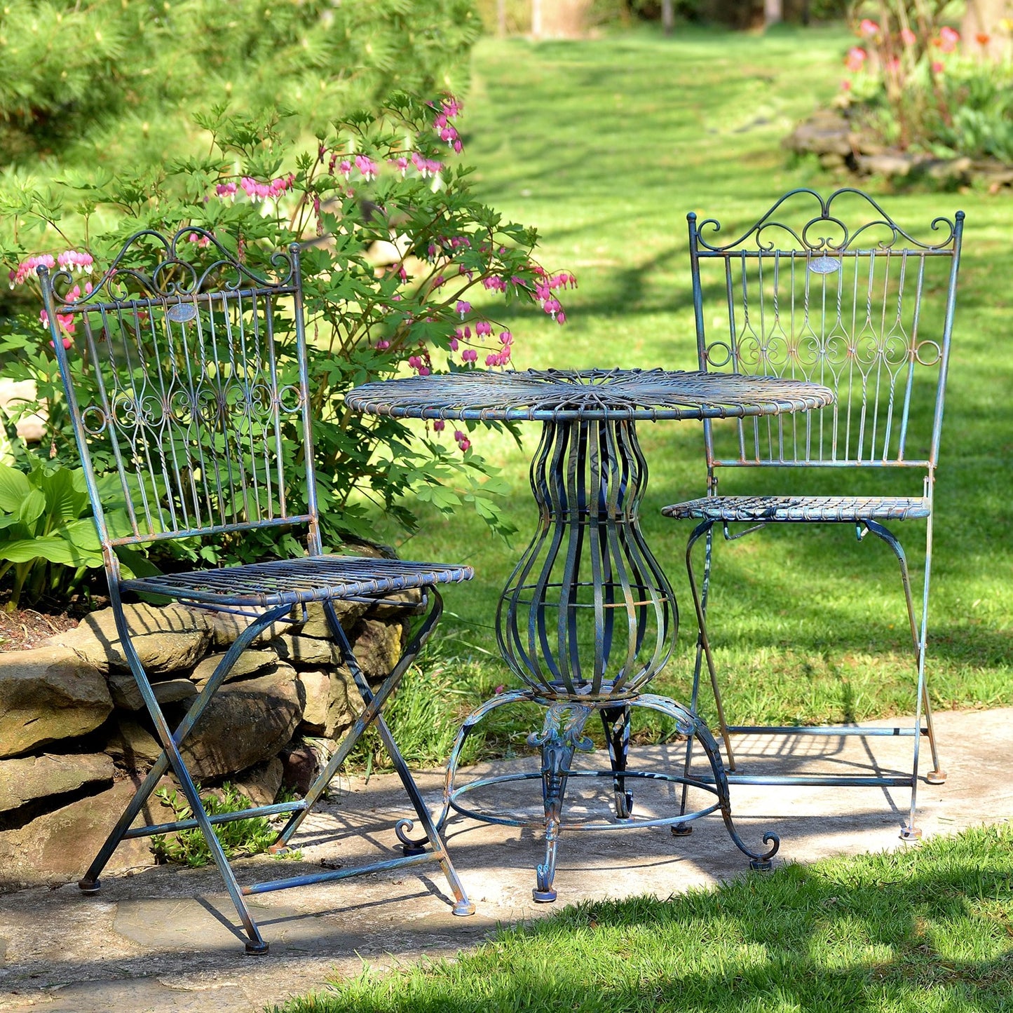 A three-piece metal garden bistro set with a round table and two matching chairs, painted in a distressed blue-green finish, set on a stone patio in a grassy garden.