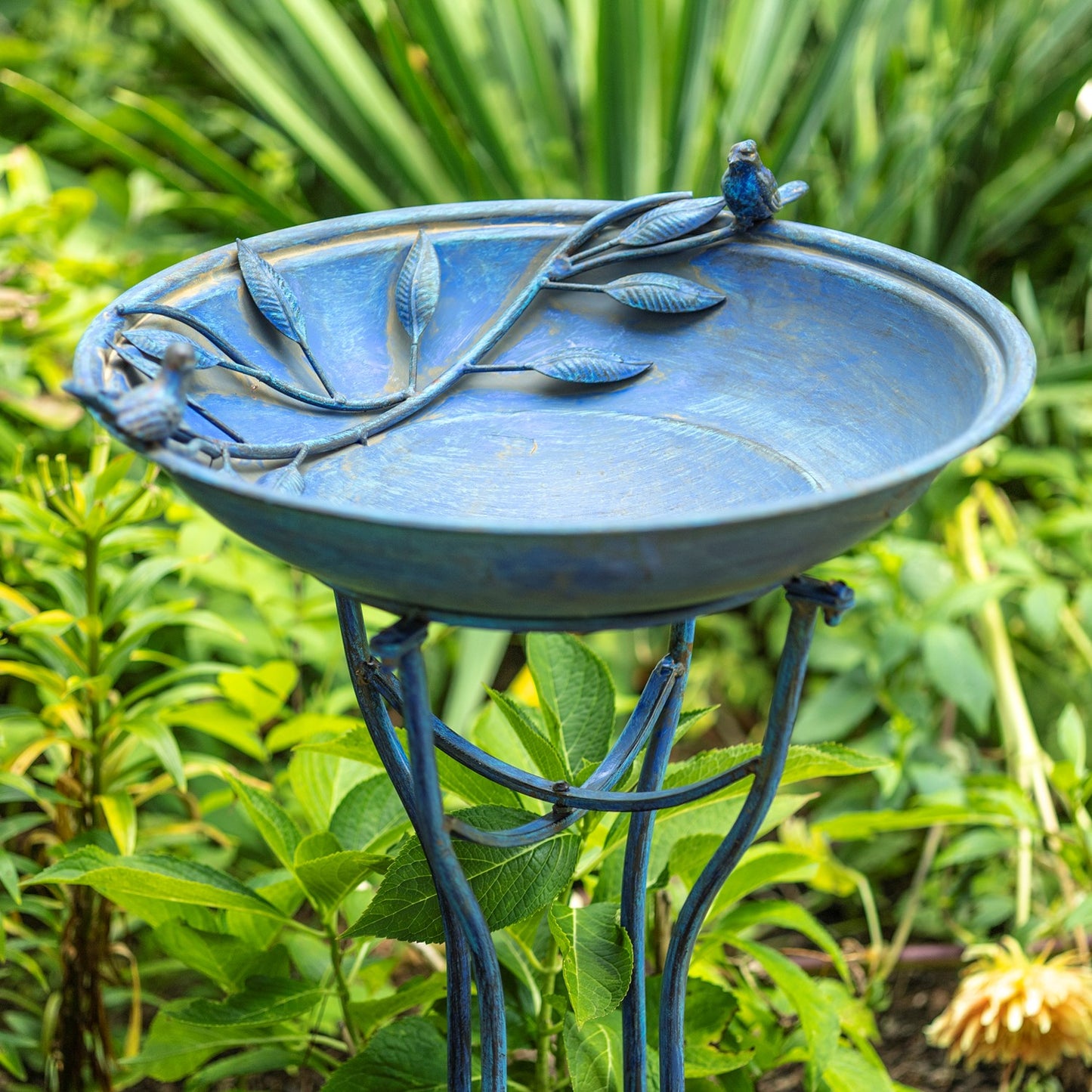 A blue metal garden birdbath with leaf and branch details on the rim and a small bird figure perched on the edge, set in a green garden.