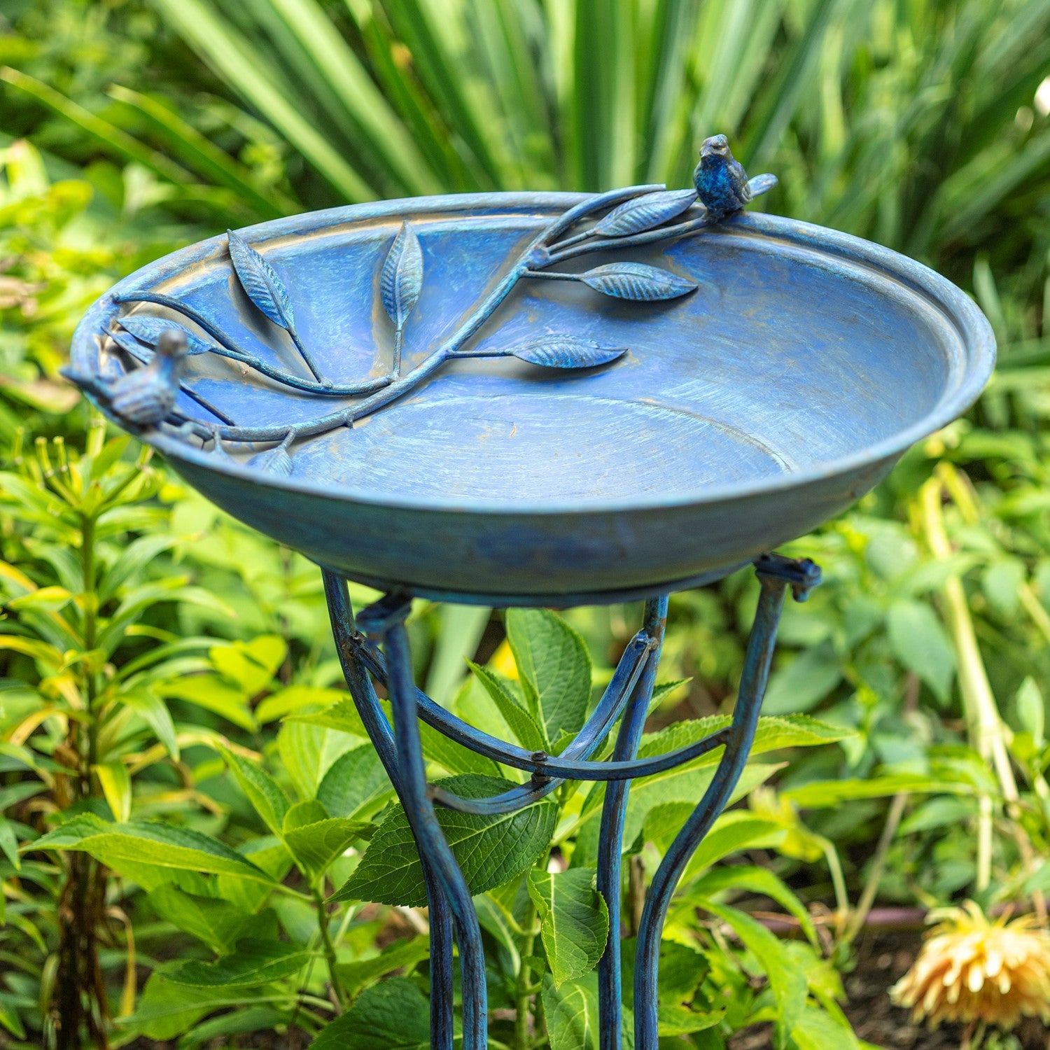 A blue metal garden birdbath with leaf and branch details on the rim and a small bird figure perched on the edge, set in a green garden.