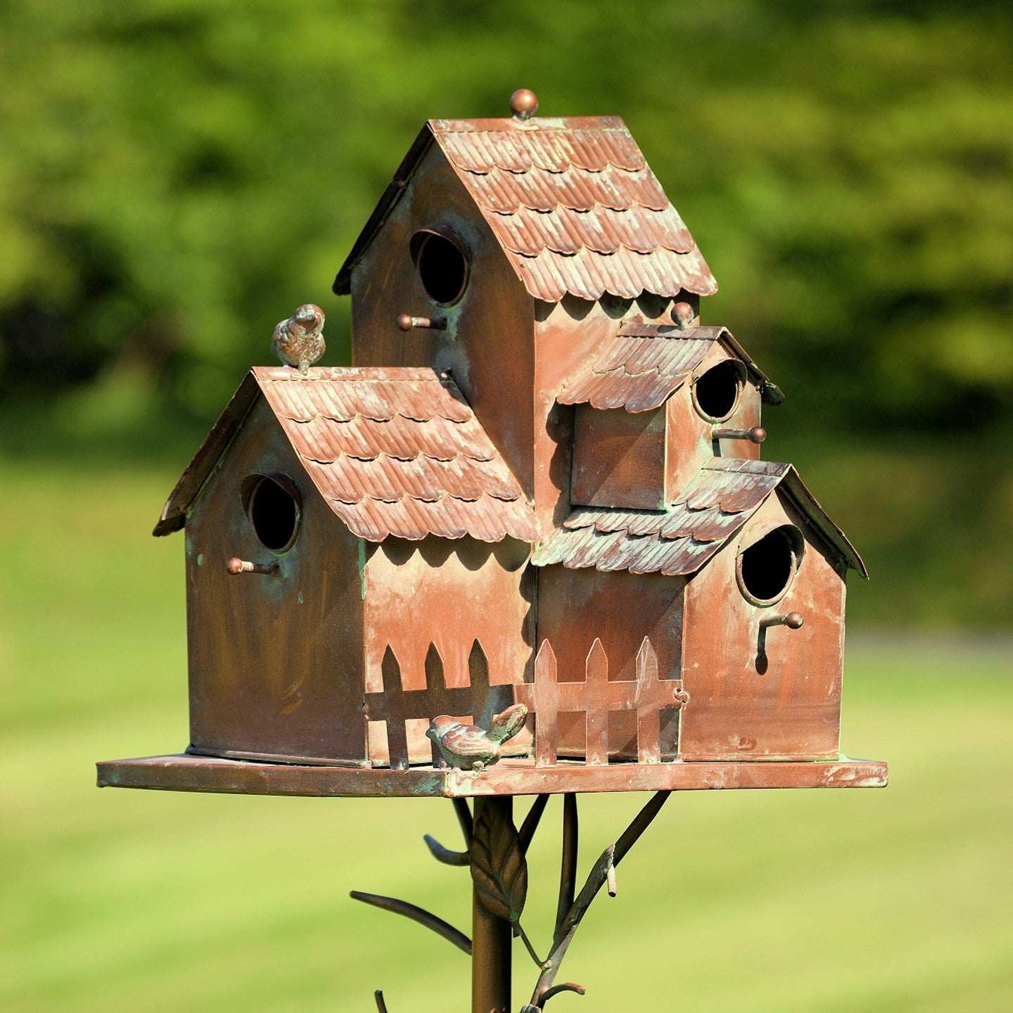 A rustic multi-tiered metal birdhouse with a weathered copper-brown patina and small bird perches, photographed outdoors against a blurred green lawn background.