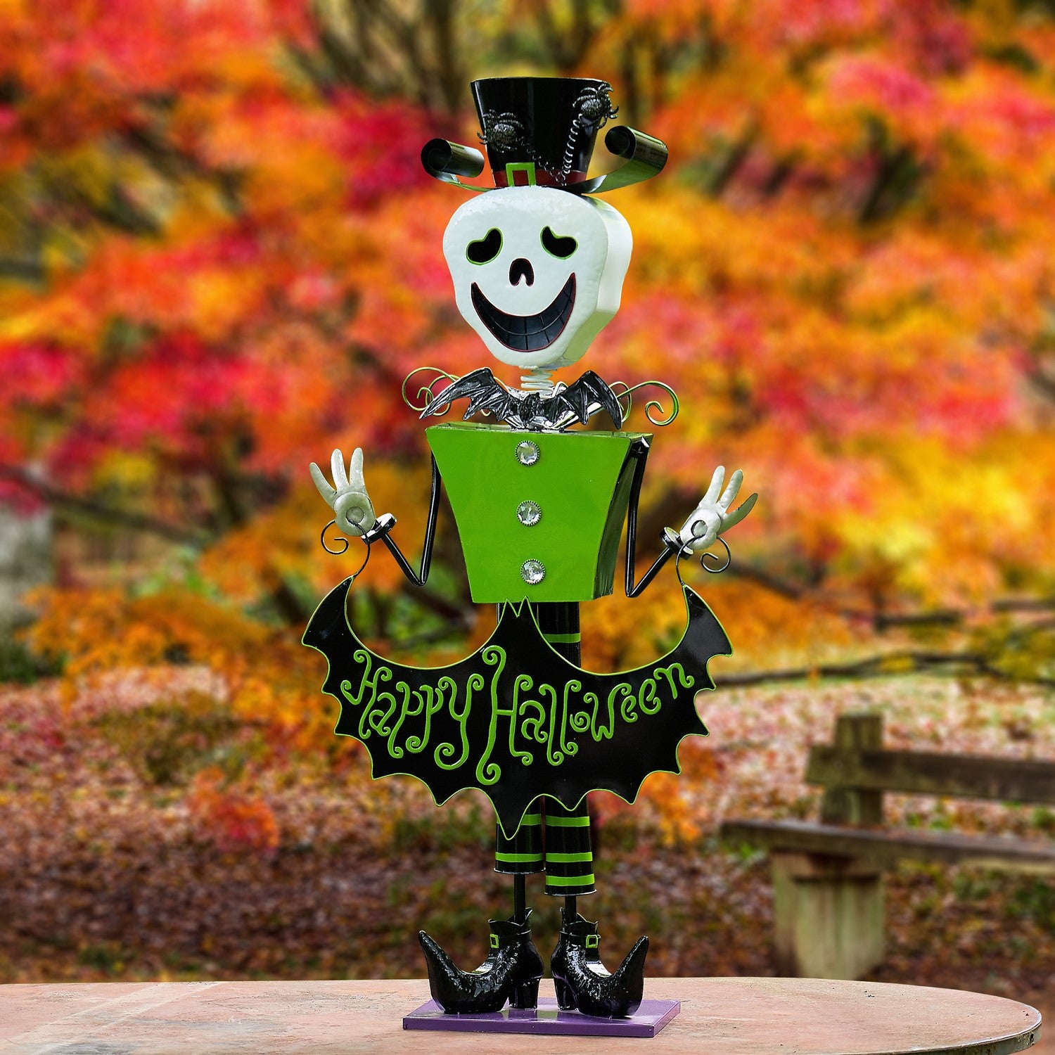 A decorative metal Halloween figure styled like a skeleton character with a black top hat, white face, green torso, black bat-wing skirt with 'Happy Halloween' text, and black shoes, photographed outdoors in front of a blurred autumn foliage background.