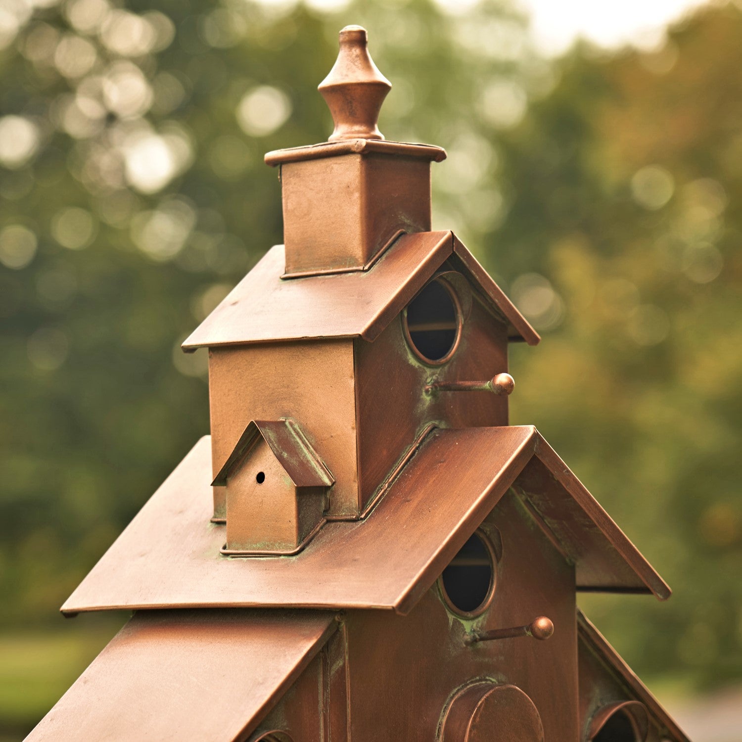 A close-up of a decorative copper birdhouse with a multi-tiered roof and small perched detail, photographed outdoors against a blurred green background.