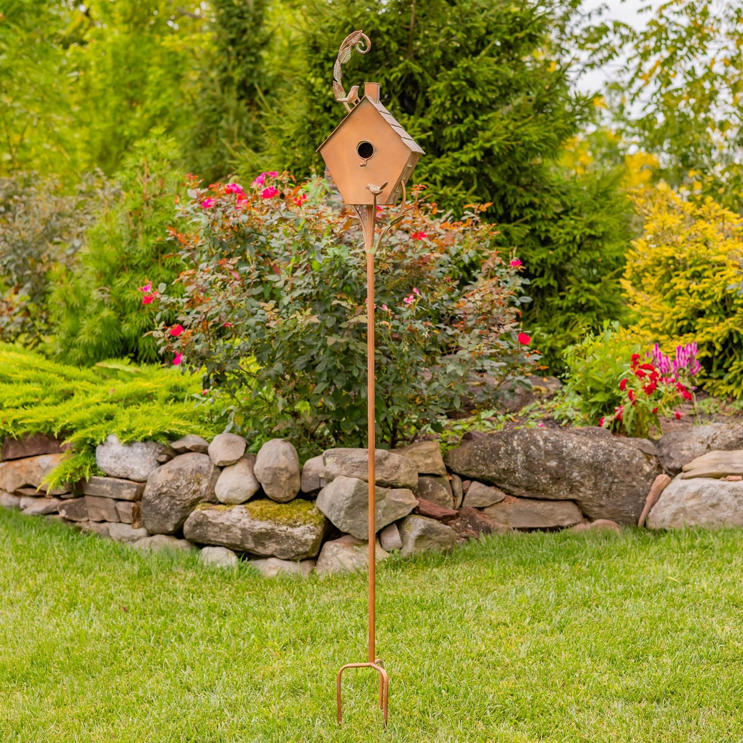 Decorative copper birdhouse with a small bird on top, set against a garden background with flowers and trees