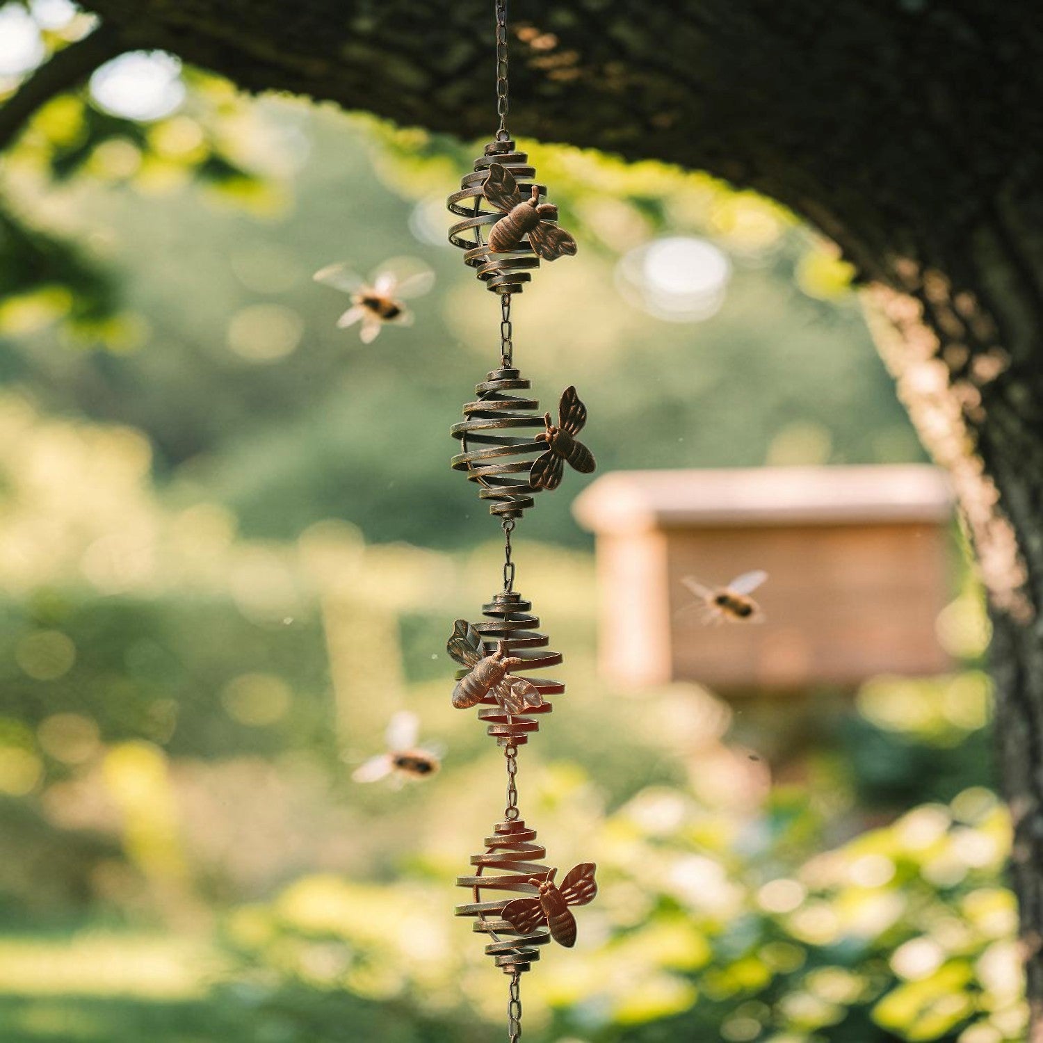 A hanging wind spinner with stacked wood discs, pinecones, and butterfly and bee figures, suspended outdoors under a tree branch with a blurred garden background.