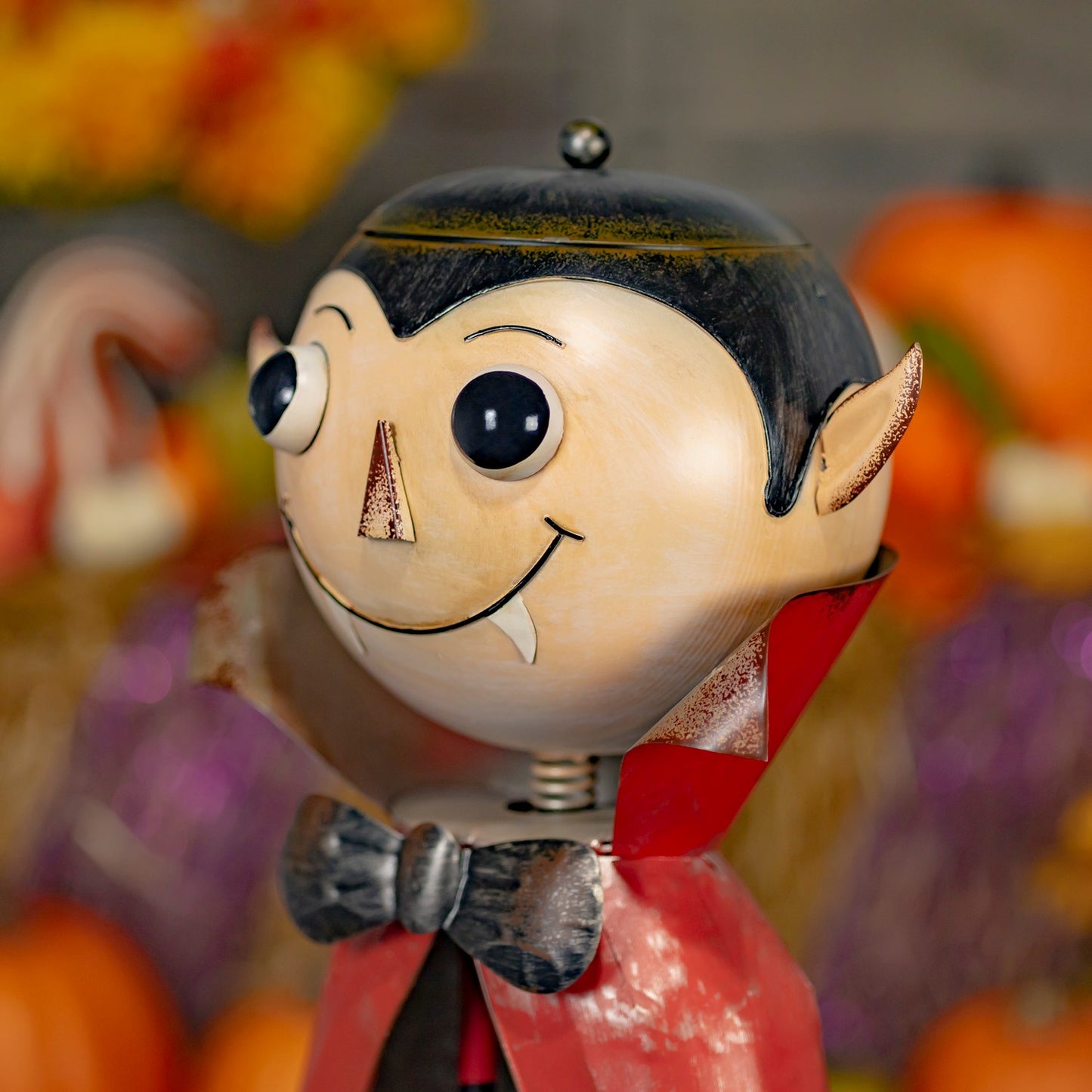 A close-up of a decorative vampire-shaped object with a beige face, black hair, pointed ears, a red cape, and a black bow tie, set against a blurred autumnal background with pumpkins and colorful shapes.