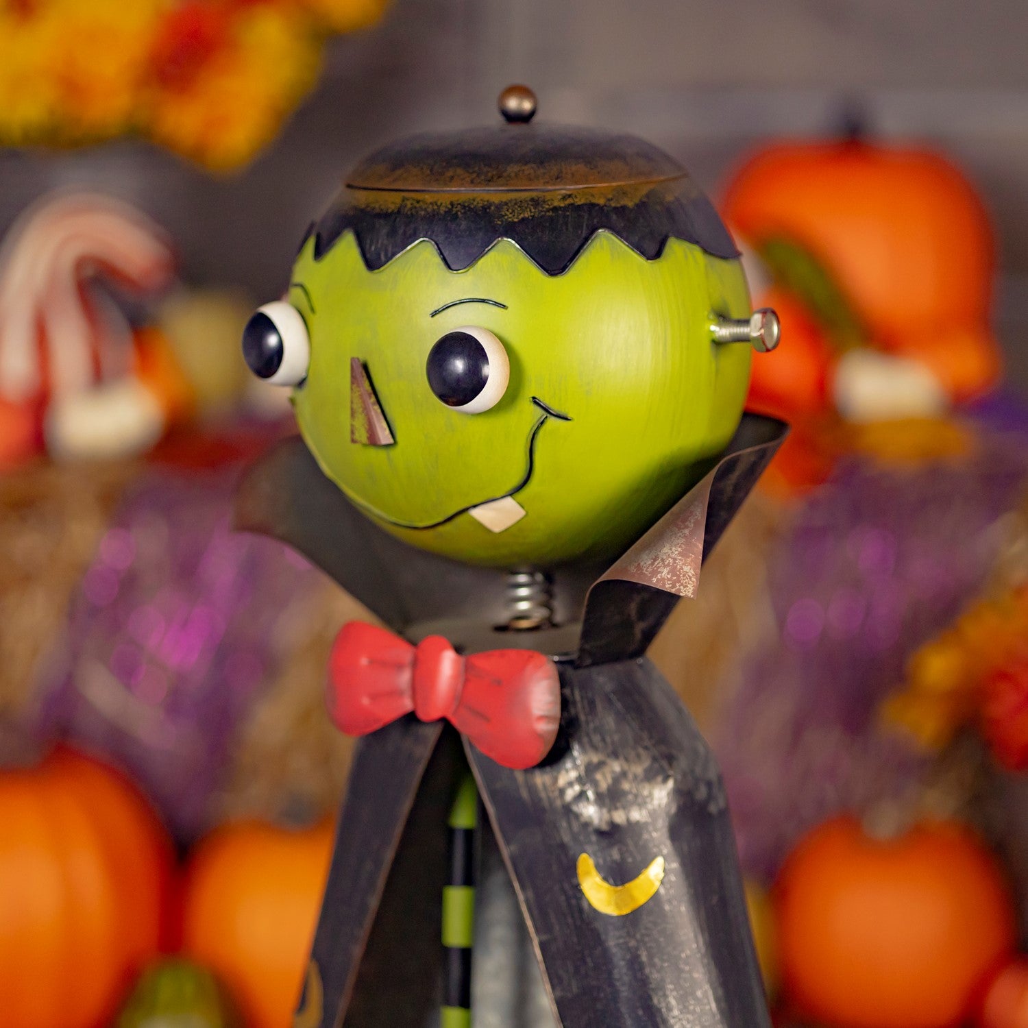 A Halloween-themed metal wind spinner toy shaped like Frankenstein's monster, featuring a lime green head, black hair, a red bow tie, and a black cape, set against a blurred autumn background with pumpkins and fall foliage.