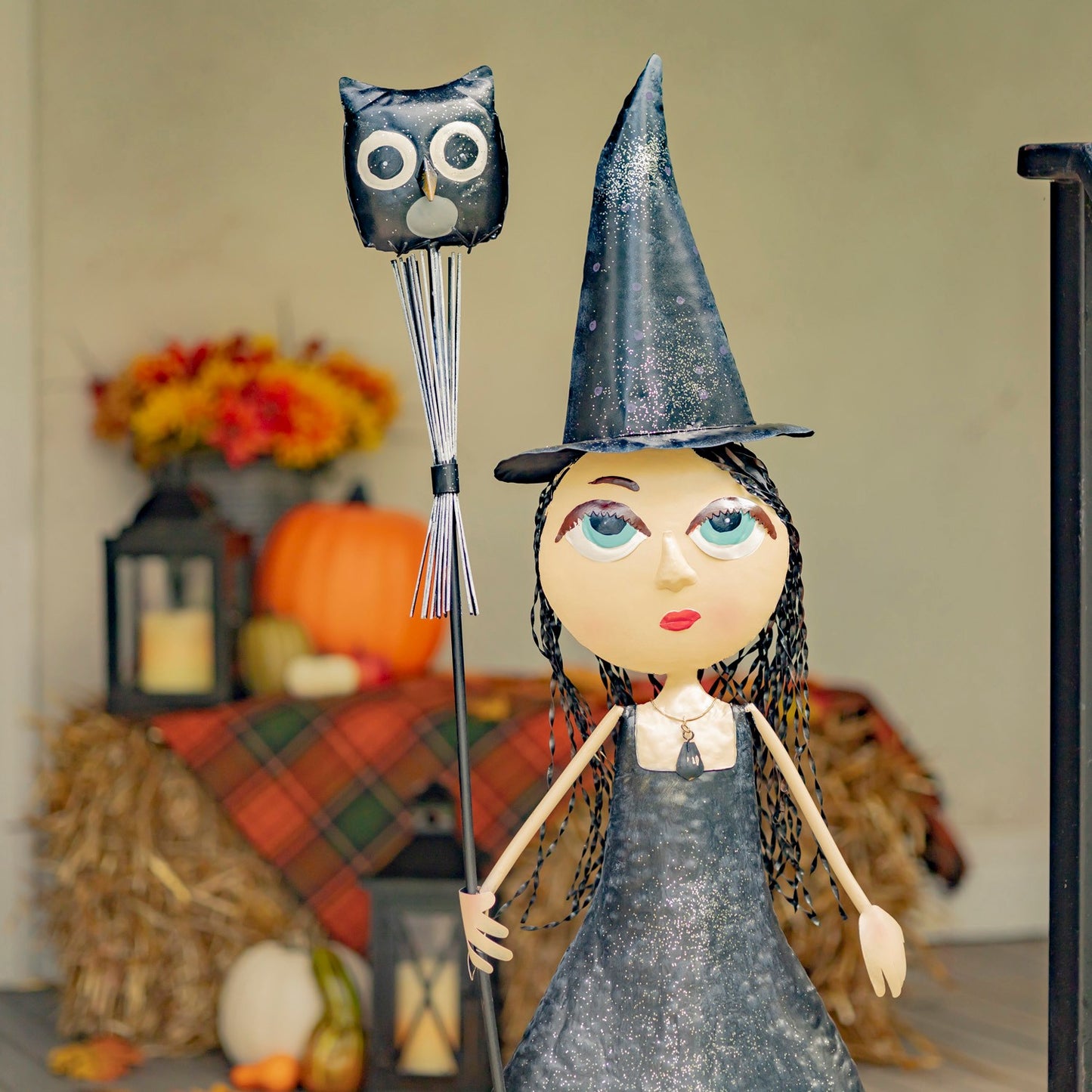 A Halloween witch doll with a glittery black hat and dress, holding a black owl-shaped wand, displayed in a fall-themed decorative setting with pumpkins, autumn leaves, and hay.