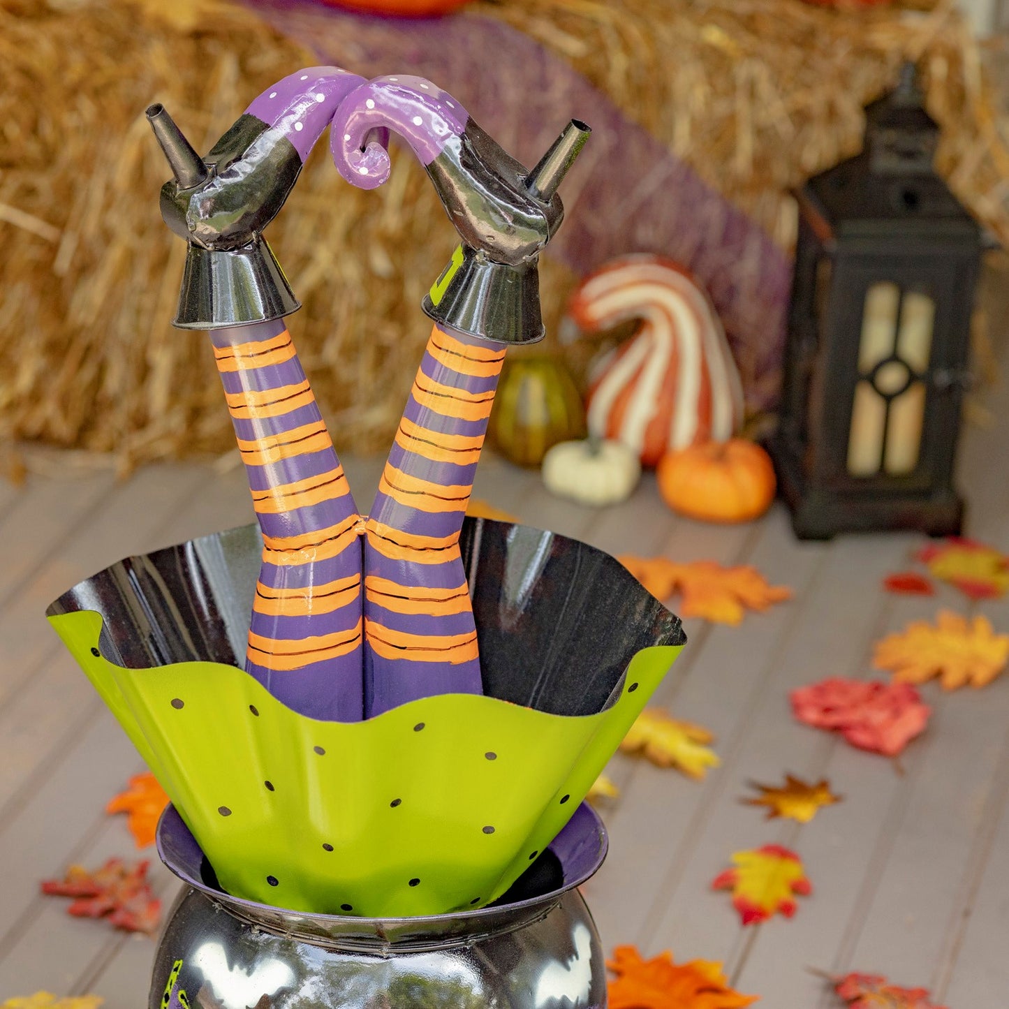 Halloween decoration of witch's legs in striped purple and orange stockings with black boots, sitting in a green polka-dot bowl inside a silver cauldron, styled with autumn leaves, pumpkins, and a lantern on a hay bale surface.