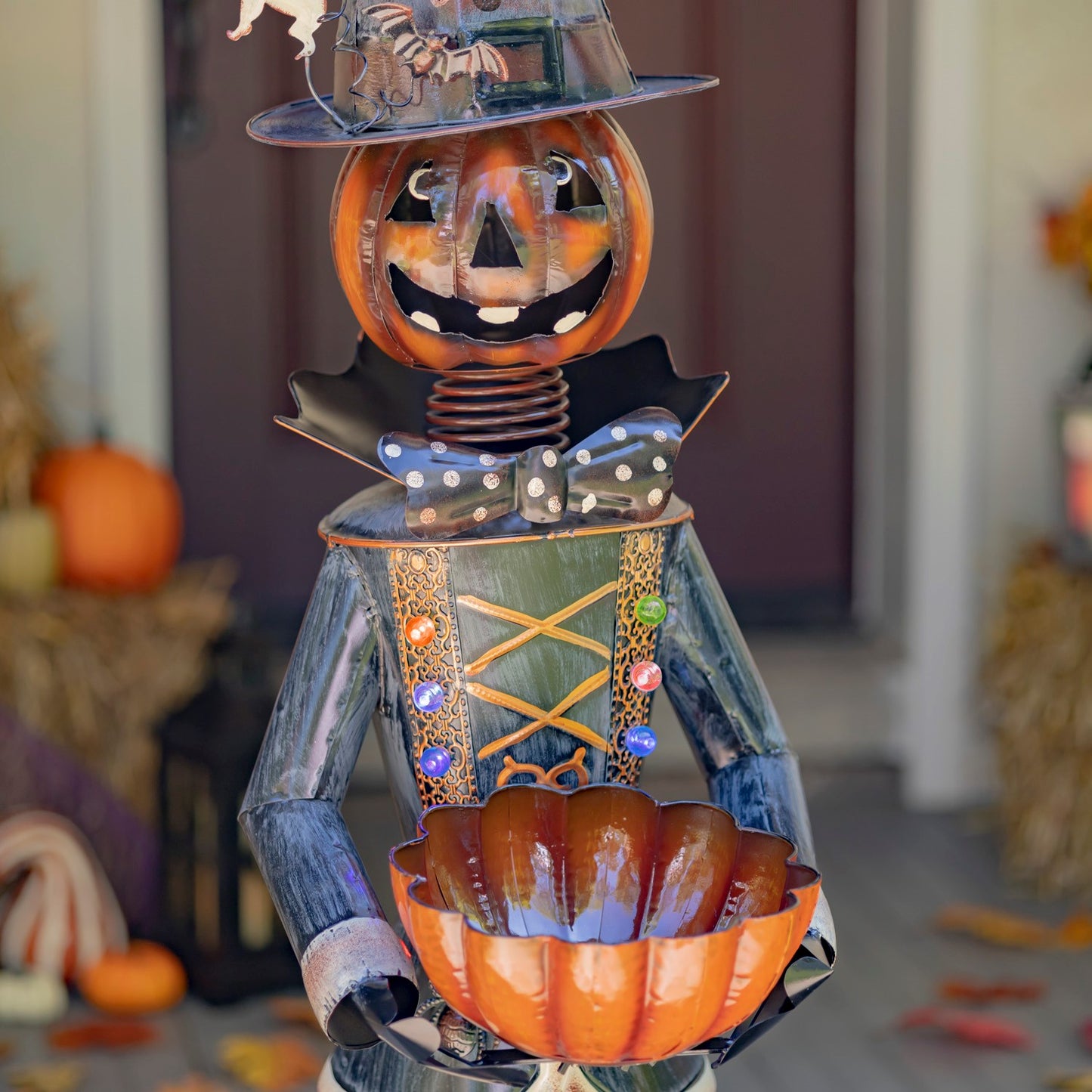 A decorative Halloween scarecrow metal garden statue holding an orange pumpkin bowl, standing on a front porch with autumn decorations in the background.