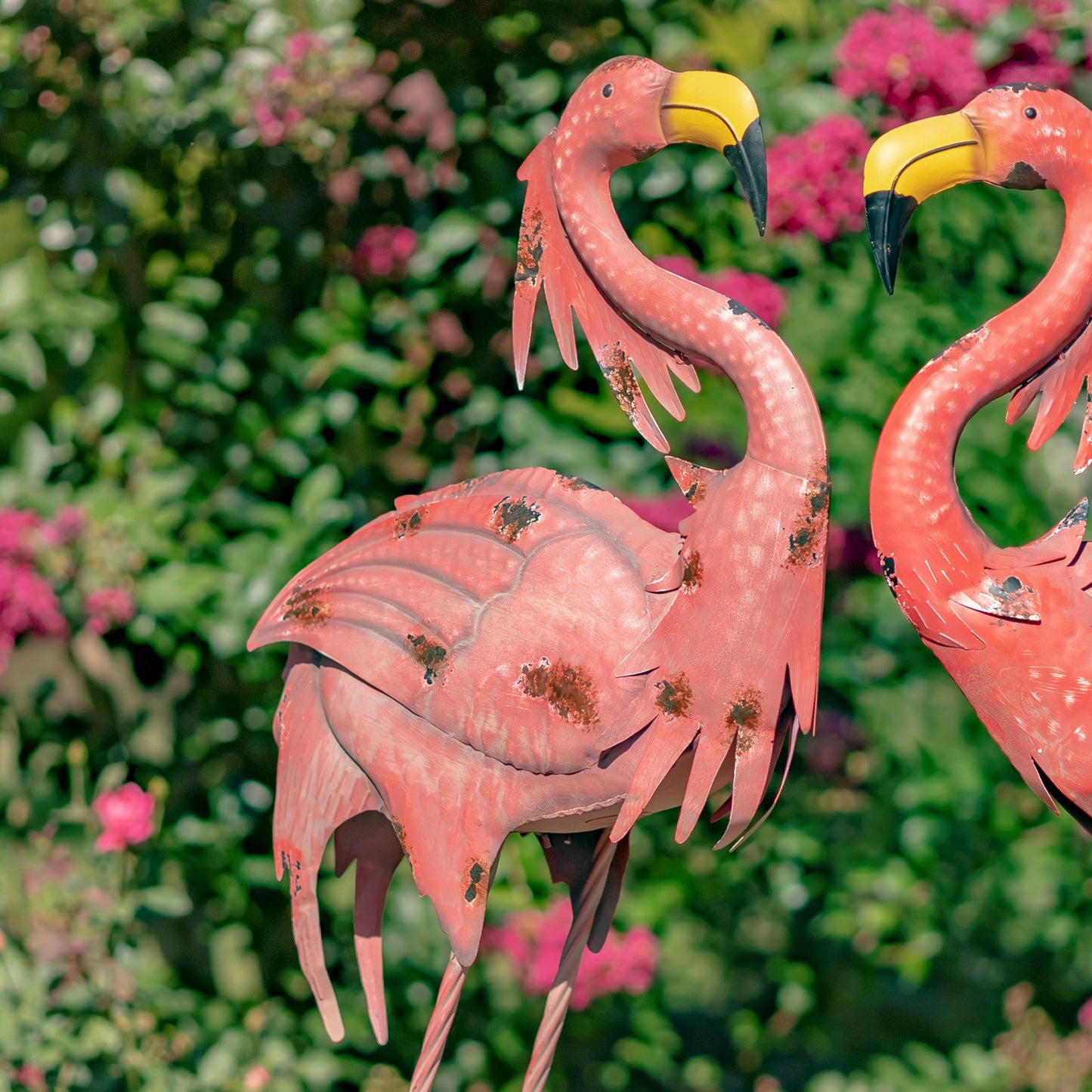 A pink metal flamingo garden statue with a yellow beak, shown outdoors against a blurred green garden background with pink flowers. A second flamingo statue is partially visible on the right.