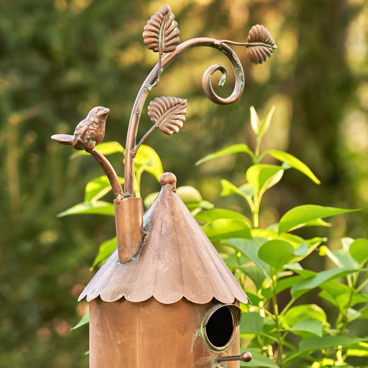 A decorative copper birdhouse with a pointed roof, scalloped edge, and a small bird perched on a leafy metal branch extending from the top, photographed outdoors against blurred green foliage.
