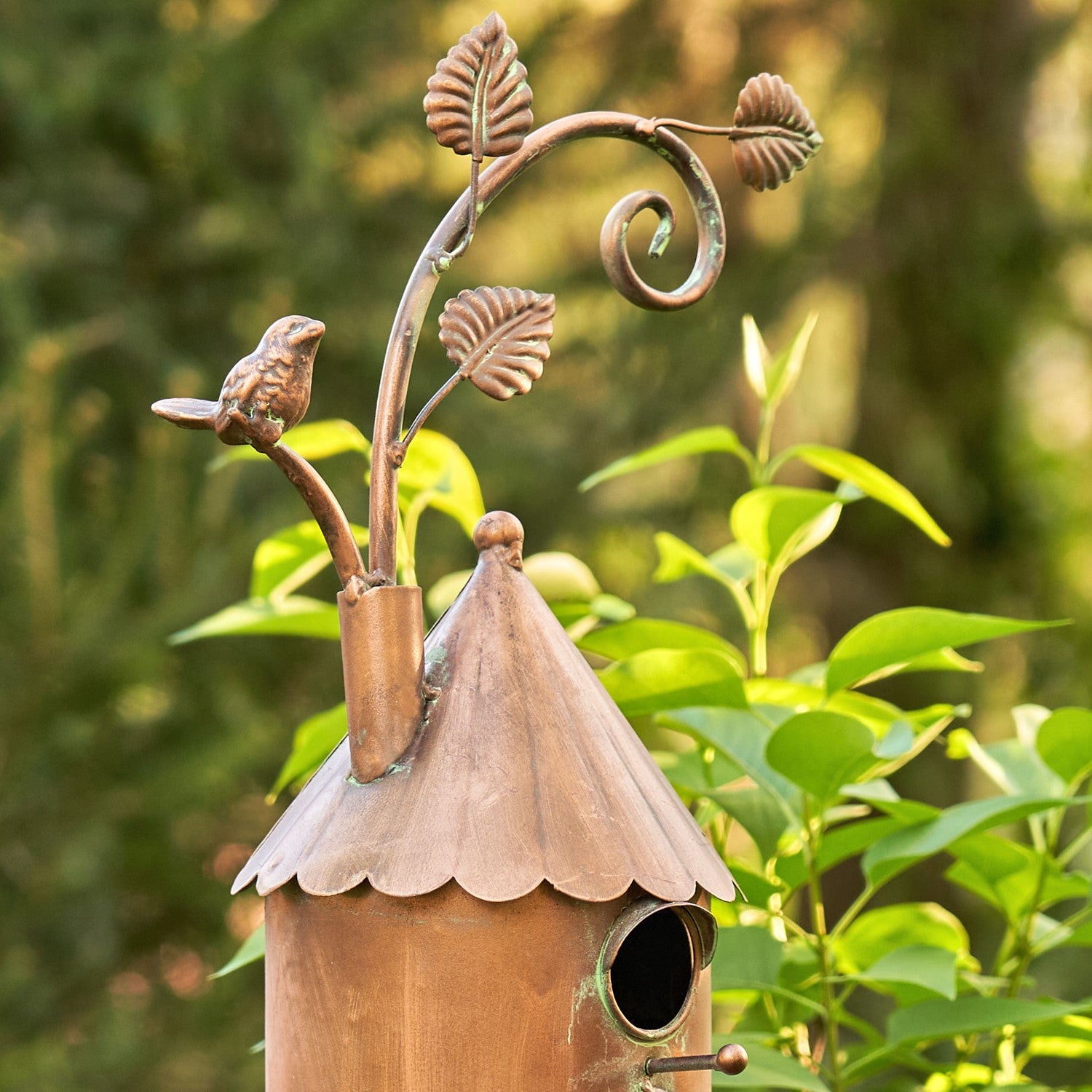 A decorative copper birdhouse with a pointed roof, scalloped edge, and a small bird perched on a leafy metal branch extending from the top, photographed outdoors against blurred green foliage.