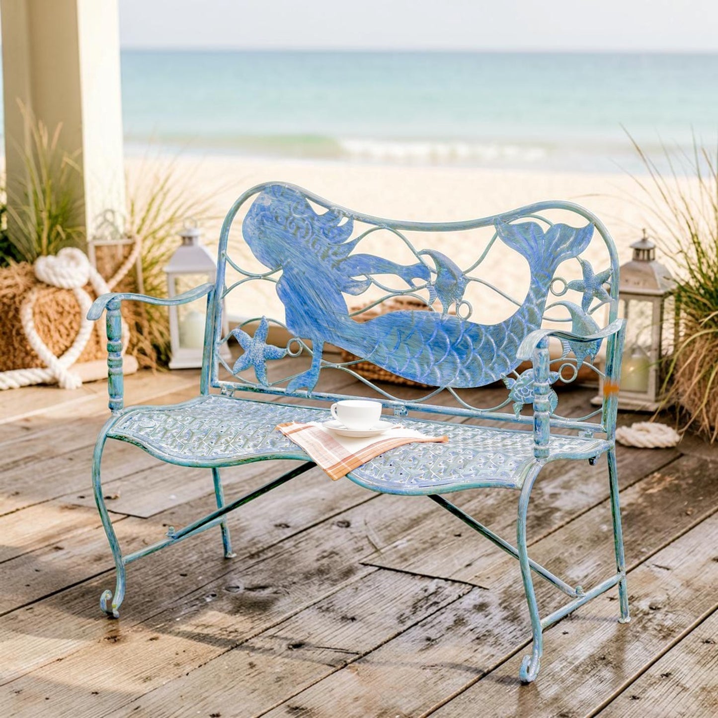 A distressed light blue metal garden bench with a mermaid and starfish design on the back, styled on a wooden deck with a beach and ocean in the background.