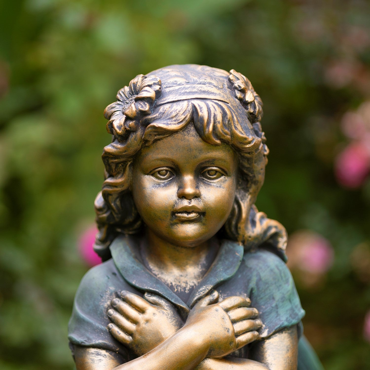 A bronze-finished garden statue of a young girl with curly hair, wearing a dark dress and standing with her arms crossed over her chest, set against a blurred green garden background.