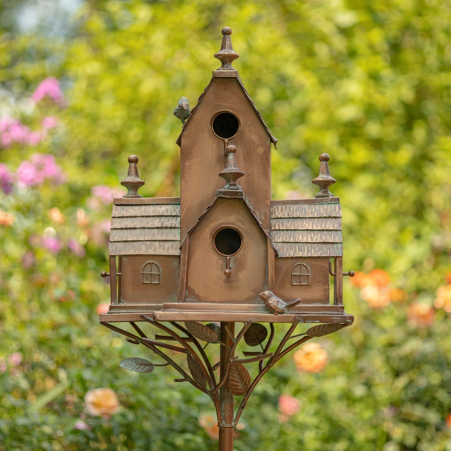 A multi-tiered birdhouse with a rustic brown and bronze finish, mounted on a decorative metal stand with leaf details, set against a blurred green garden background with pink flowers.