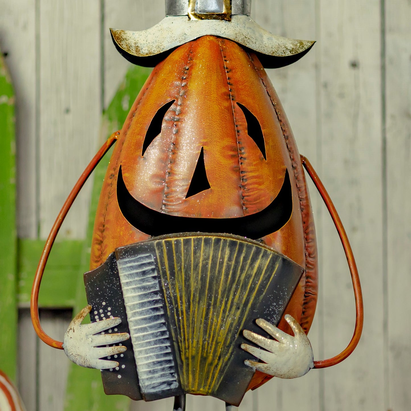A decorative metal pumpkin figure wearing a white hat and playing an accordion, photographed against a light wood plank background.