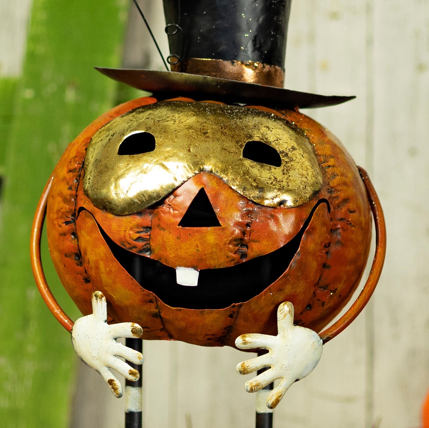 A decorative metal jack-o'-lantern Halloween decoration with a gold mask, black top hat, orange body, and white hands, photographed against a blurred green and white background.