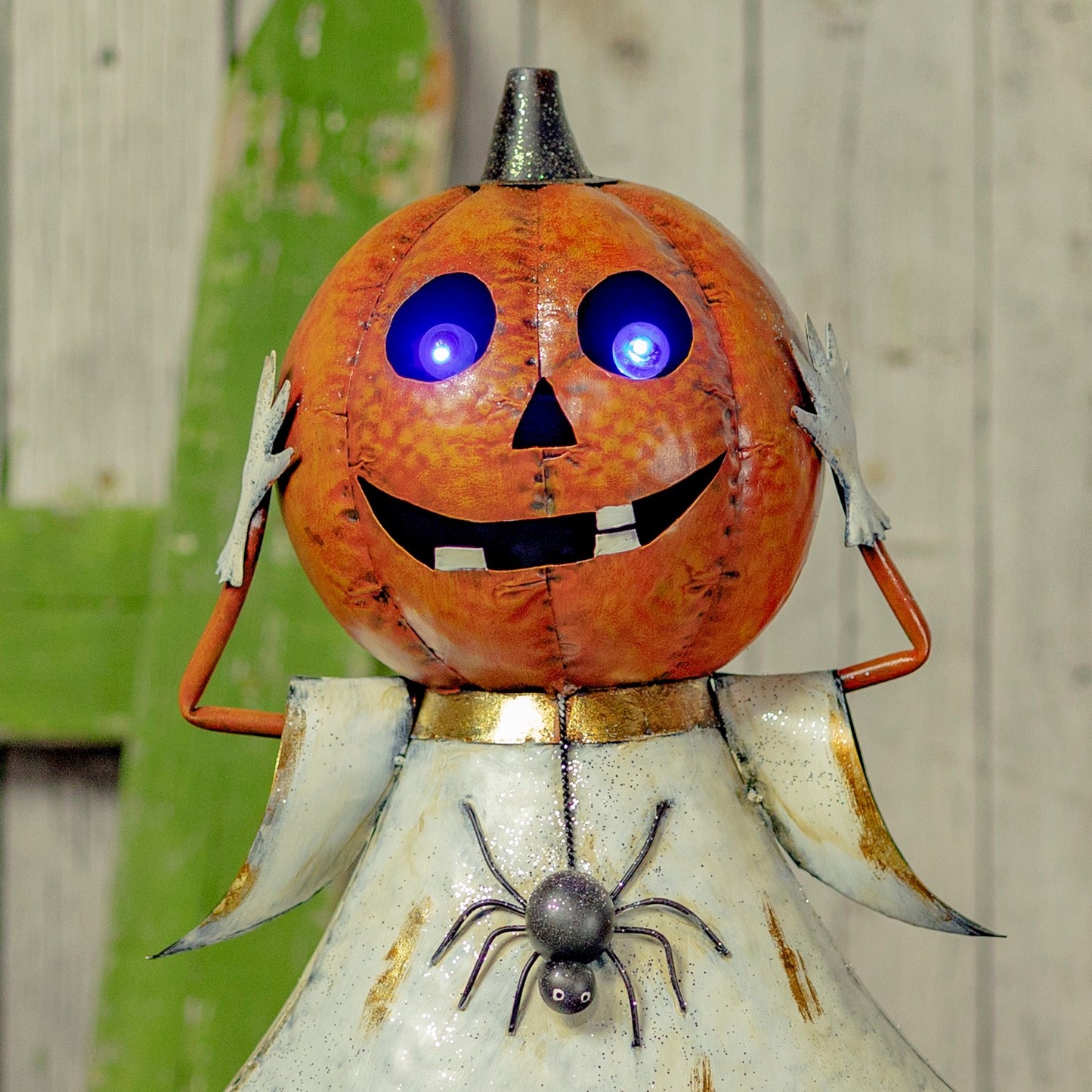 A Halloween metal pumpkin jack-o'-lantern decoration with glowing blue LED eyes, a stitched orange face, and a distressed white robe with a hanging spider detail, set against a rustic wooden background.