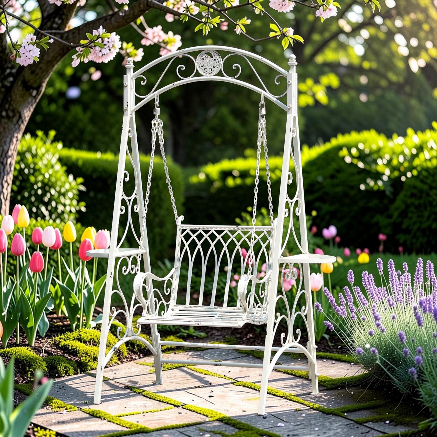 A white wrought-iron garden swing bench placed on a stone path in a lush garden with pink blossoms, tulips, and lavender.