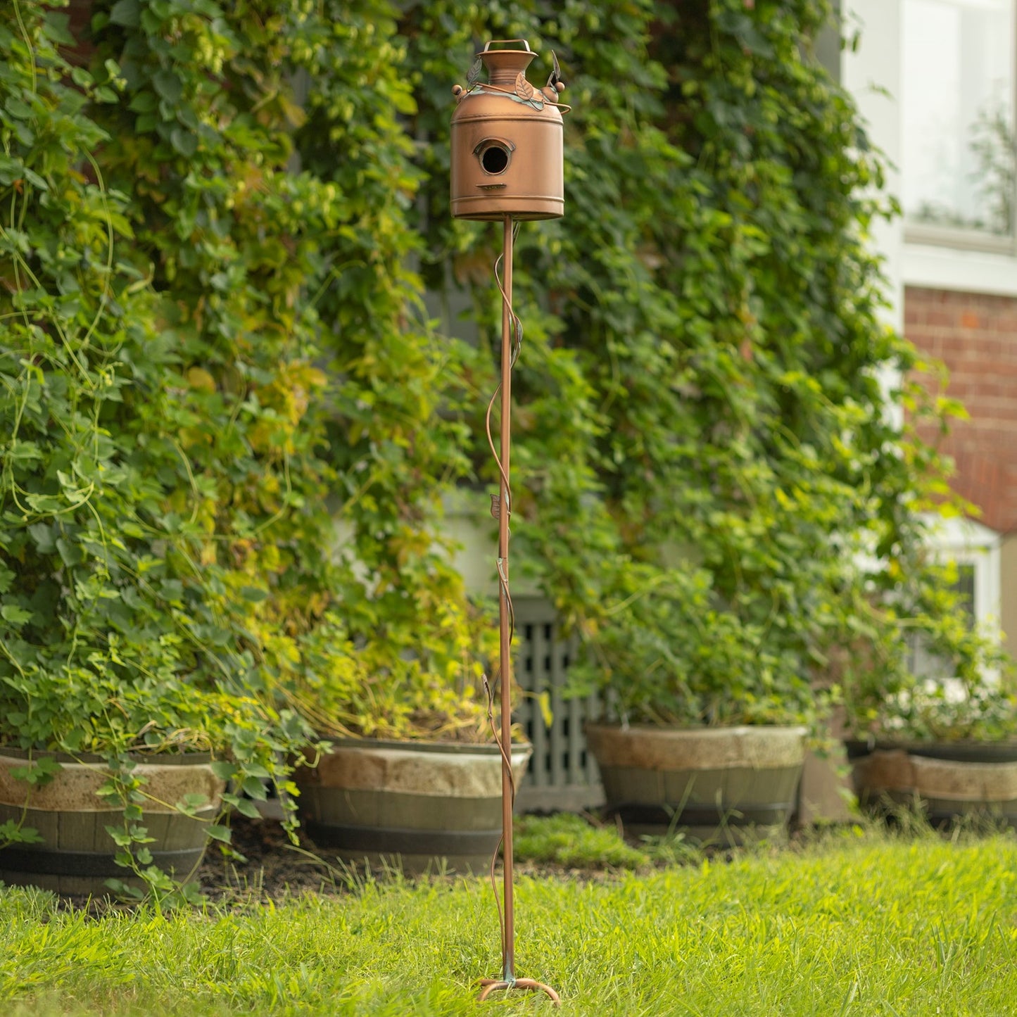 A copper birdhouse on a tall twisted metal stake, set outdoors on a green lawn in front of a leafy green garden wall.