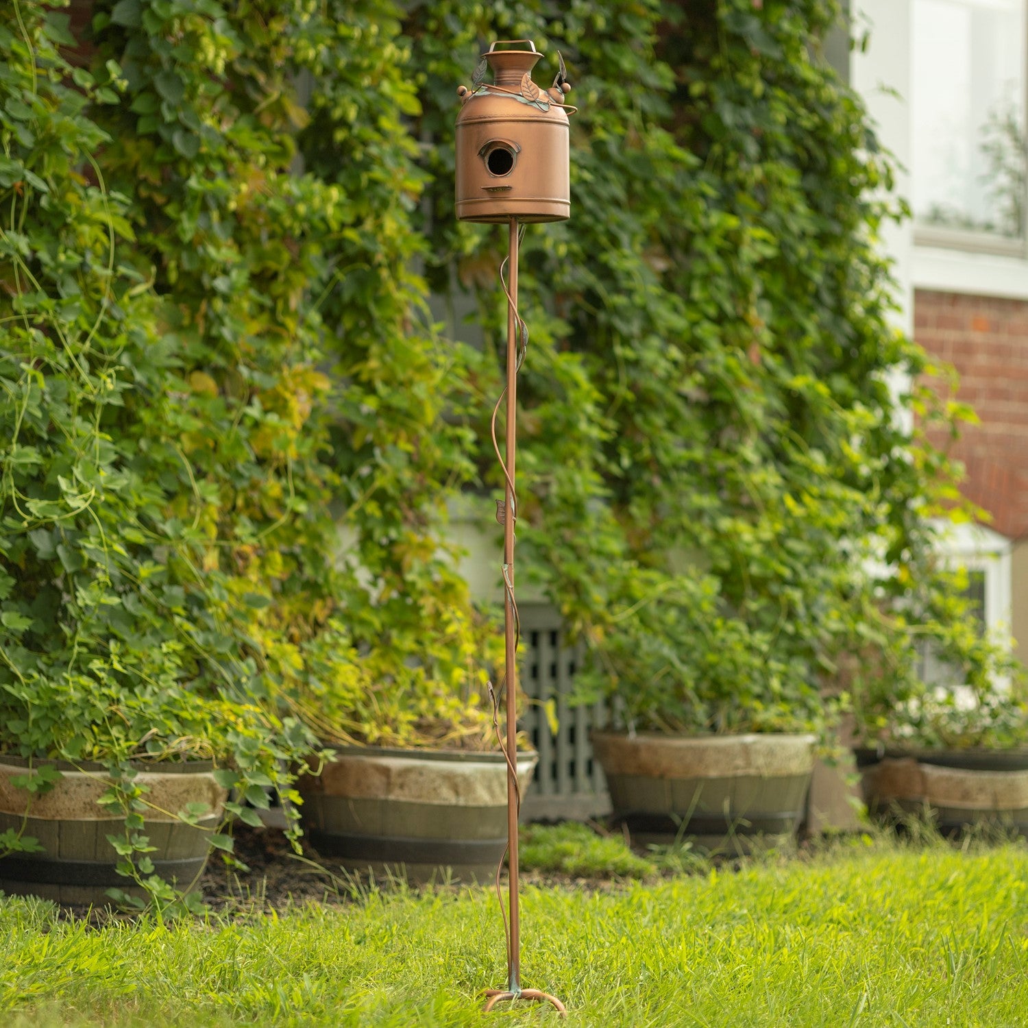 A copper birdhouse on a tall twisted metal stake, set outdoors on a green lawn in front of a leafy green garden wall.