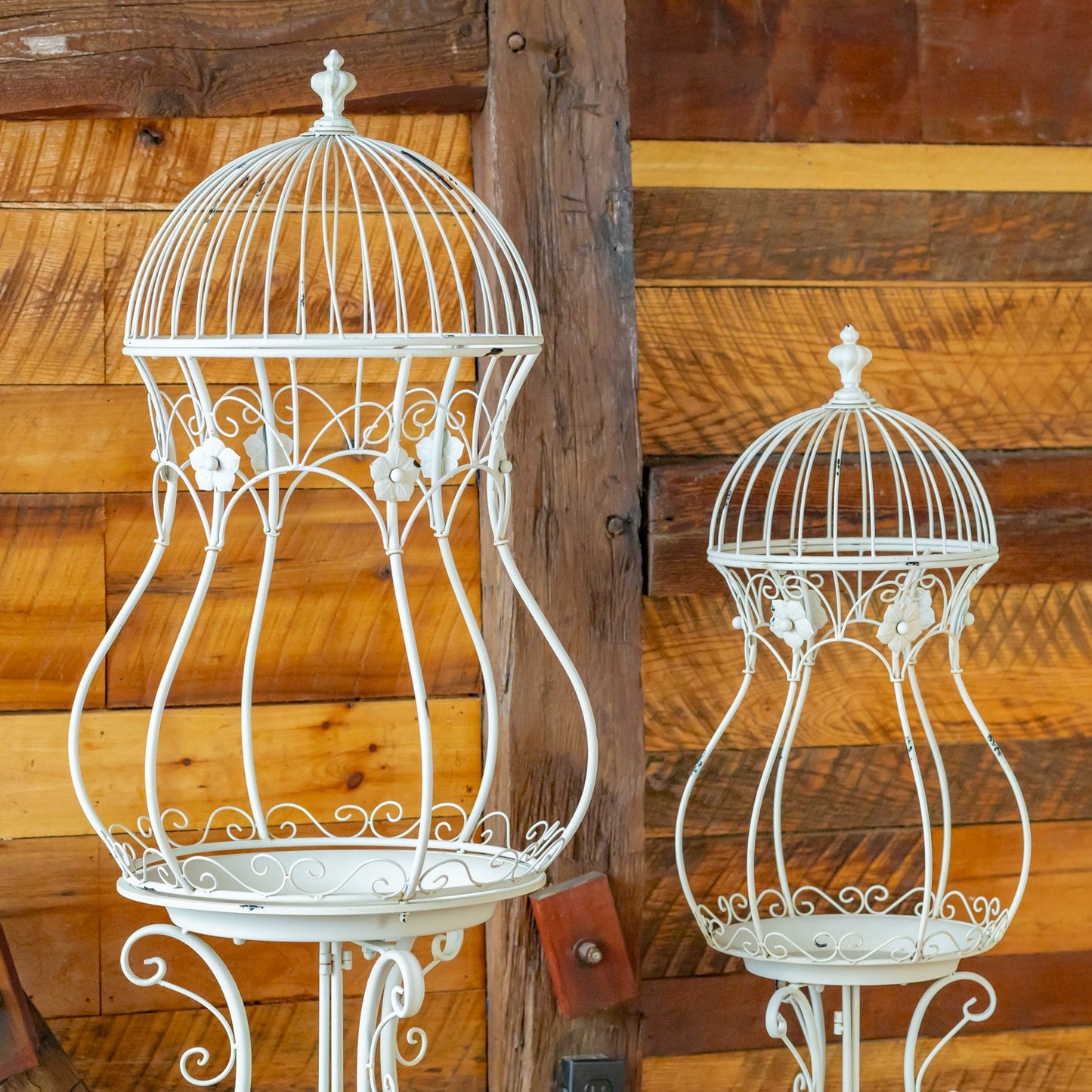 Two white metal birdcage-style decorative stands displayed against a rustic wooden plank wall, with one larger stand on the left and one smaller stand on the right.