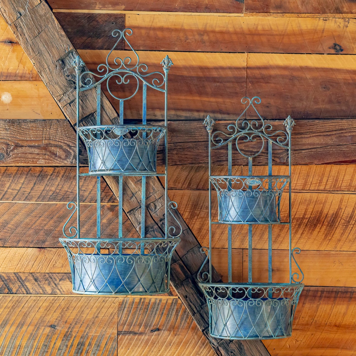 Two rustic metal tiered plant hangers with ornate scrollwork, displayed against a warm wood plank wall.