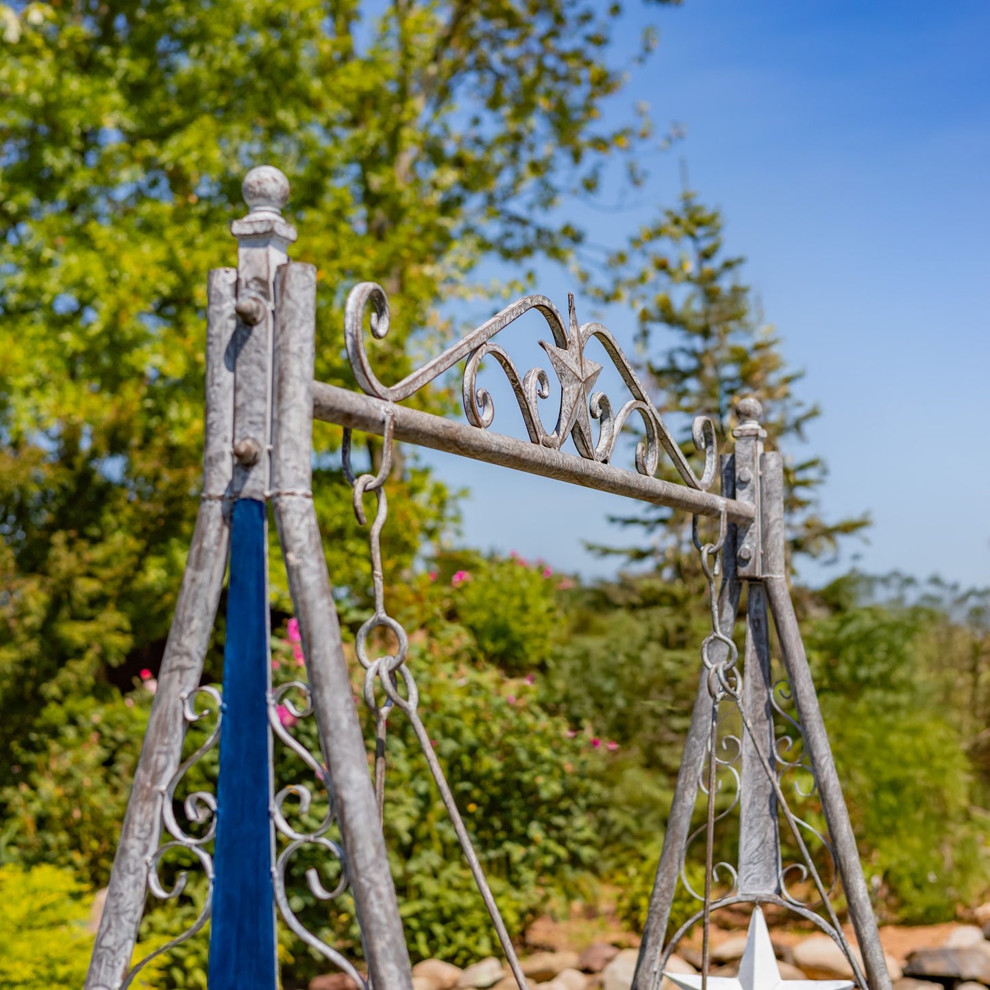 A rustic metal swing set with weathered gray and silver painted ironwork, set outdoors against a background of green trees and blue sky.