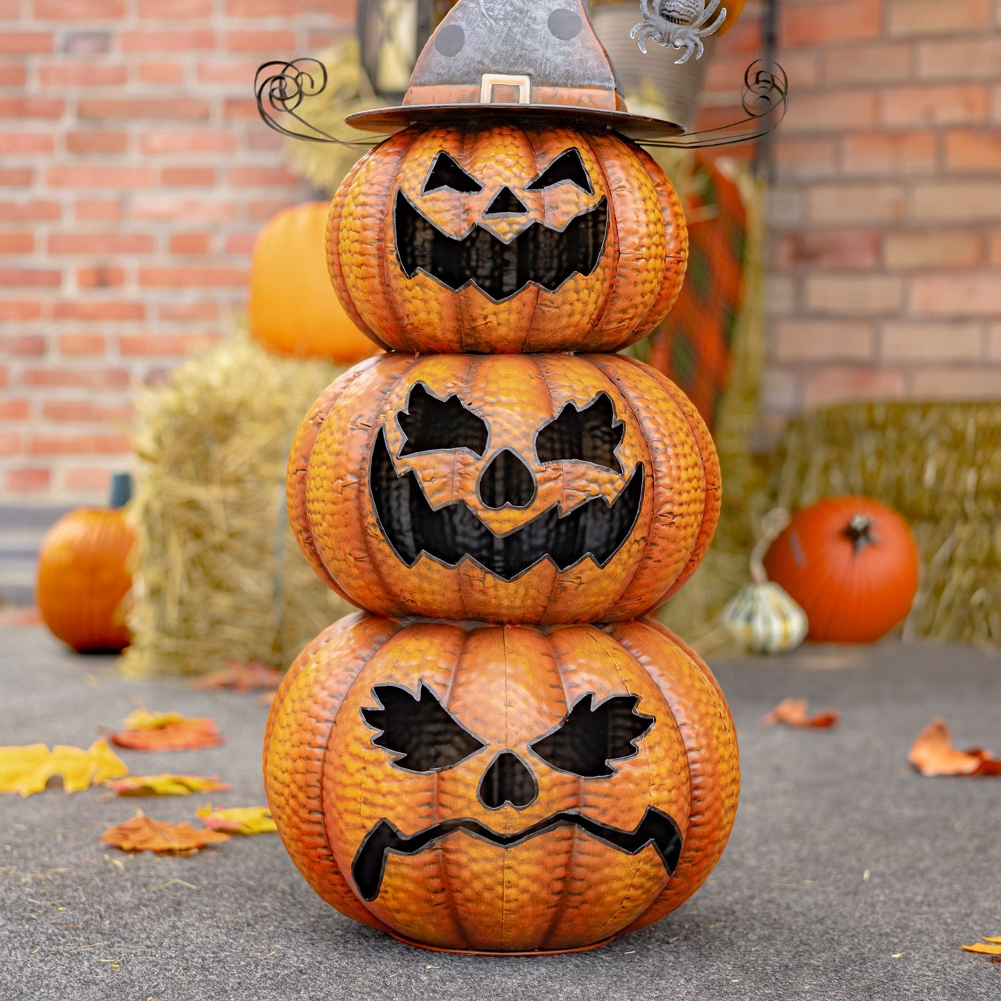 Three stacked orange jack-o'-lantern pumpkins with carved black faces, topped with a witch hat, displayed outdoors on a stone surface with a brick wall and hay bales in the background.