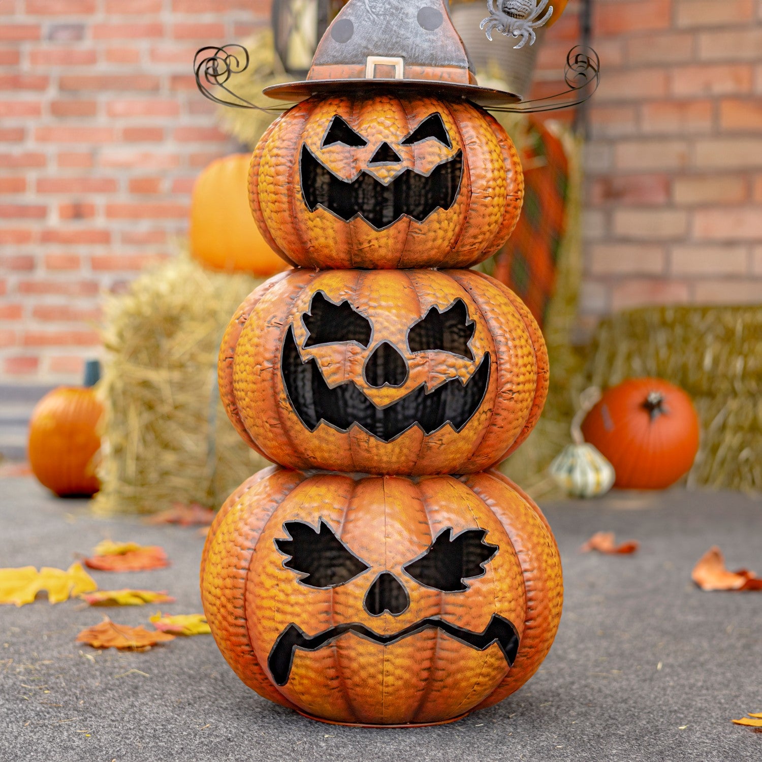 Three stacked orange jack-o'-lantern pumpkins with carved black faces, topped with a witch hat, displayed outdoors on a stone surface with a brick wall and hay bales in the background.
