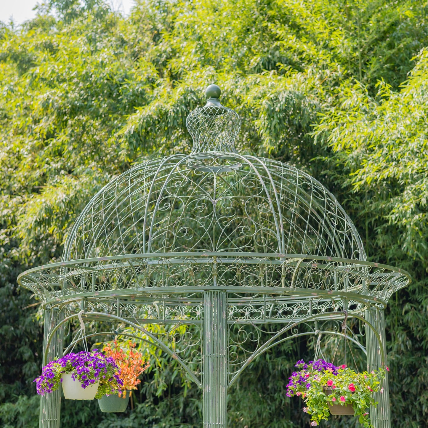 A green ornate metal gazebo with a domed roof and hanging flower baskets, set against a dense background of green trees.