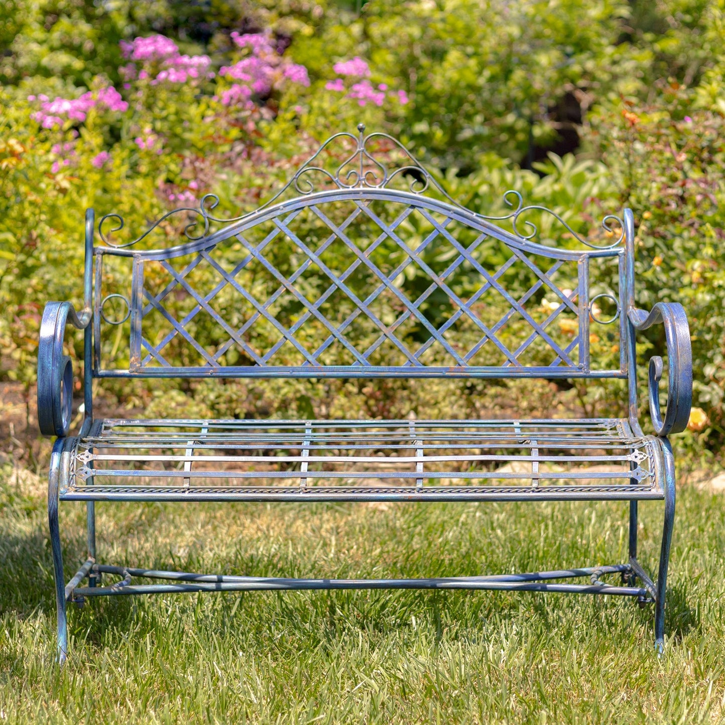 A decorative metal garden bench with an ornate blue-gray lattice backrest and curved armrests, placed on green grass in front of a blurred garden background with pink flowers.