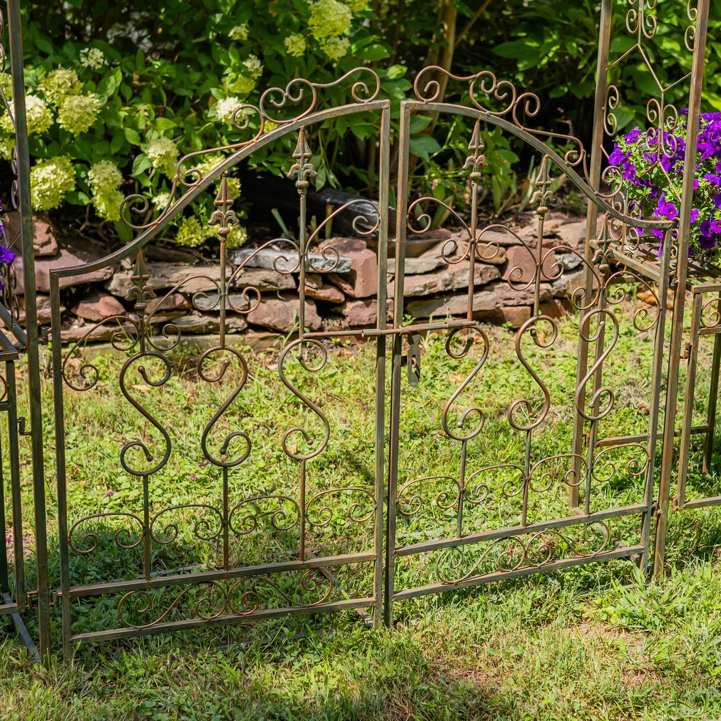 An ornate wrought iron garden gate with decorative scrollwork and a lock, set in a grassy garden with plants and flowers in the background.