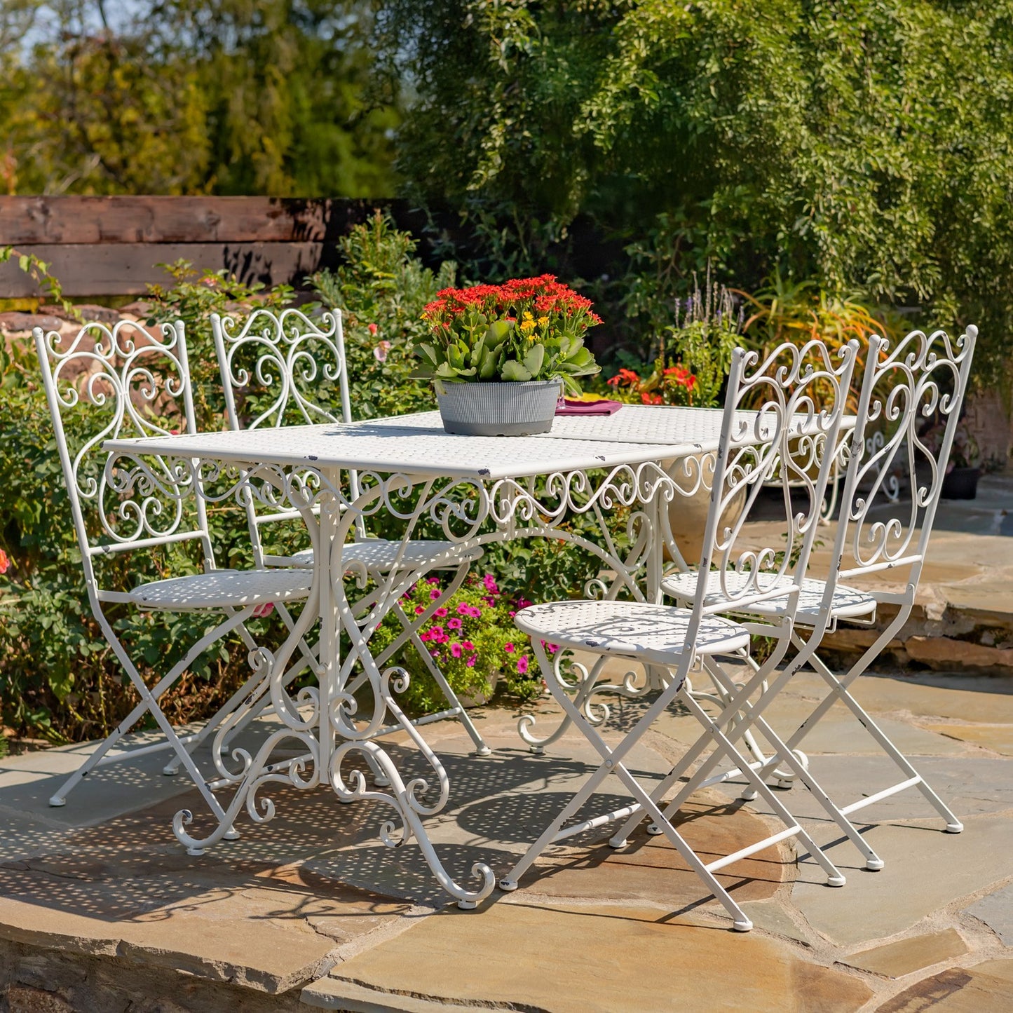 A white wrought iron outdoor bistro dining set consisting of a square table and four matching chairs, displayed on a stone patio in a lush garden setting.