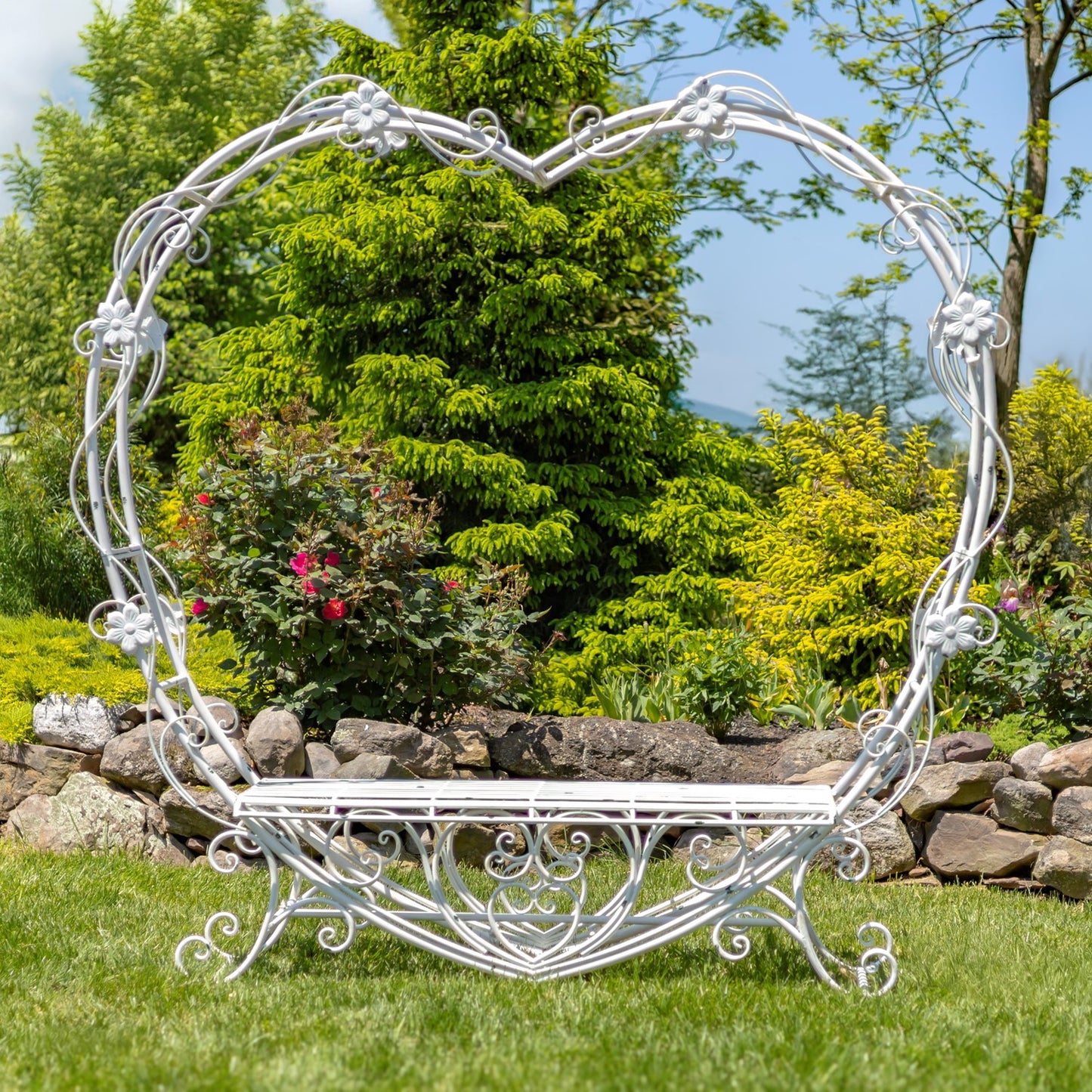 A white wrought iron garden bench shaped like an oval heart, set on a green lawn in a landscaped garden with trees, shrubs, and a stone wall.